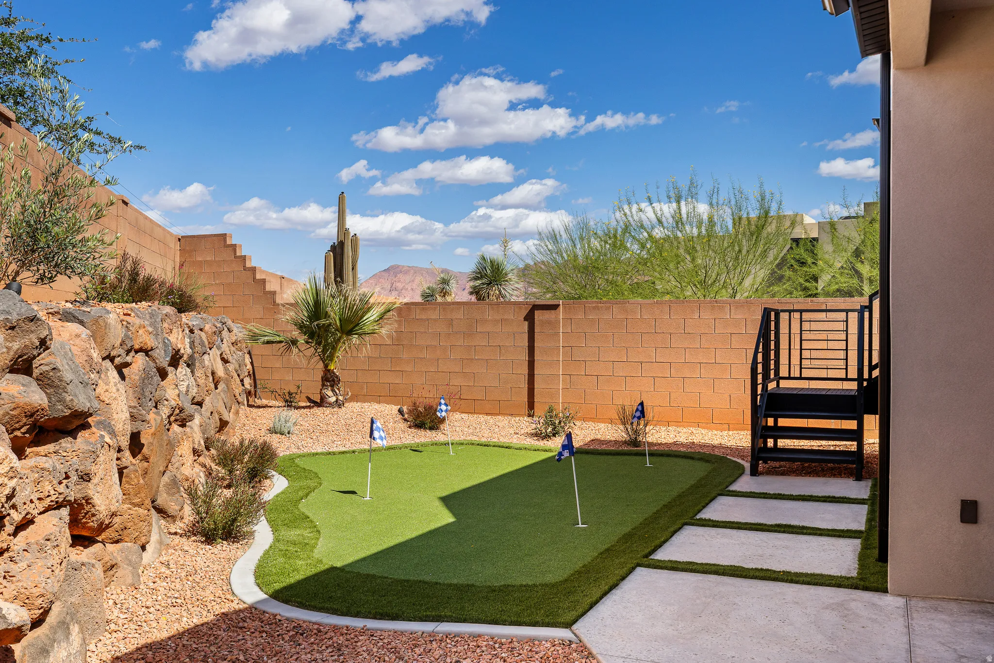 Backyard featuring a multi-hole putting green, concrete paver stepping stones, a black metal staircase, and a rock retaining wall