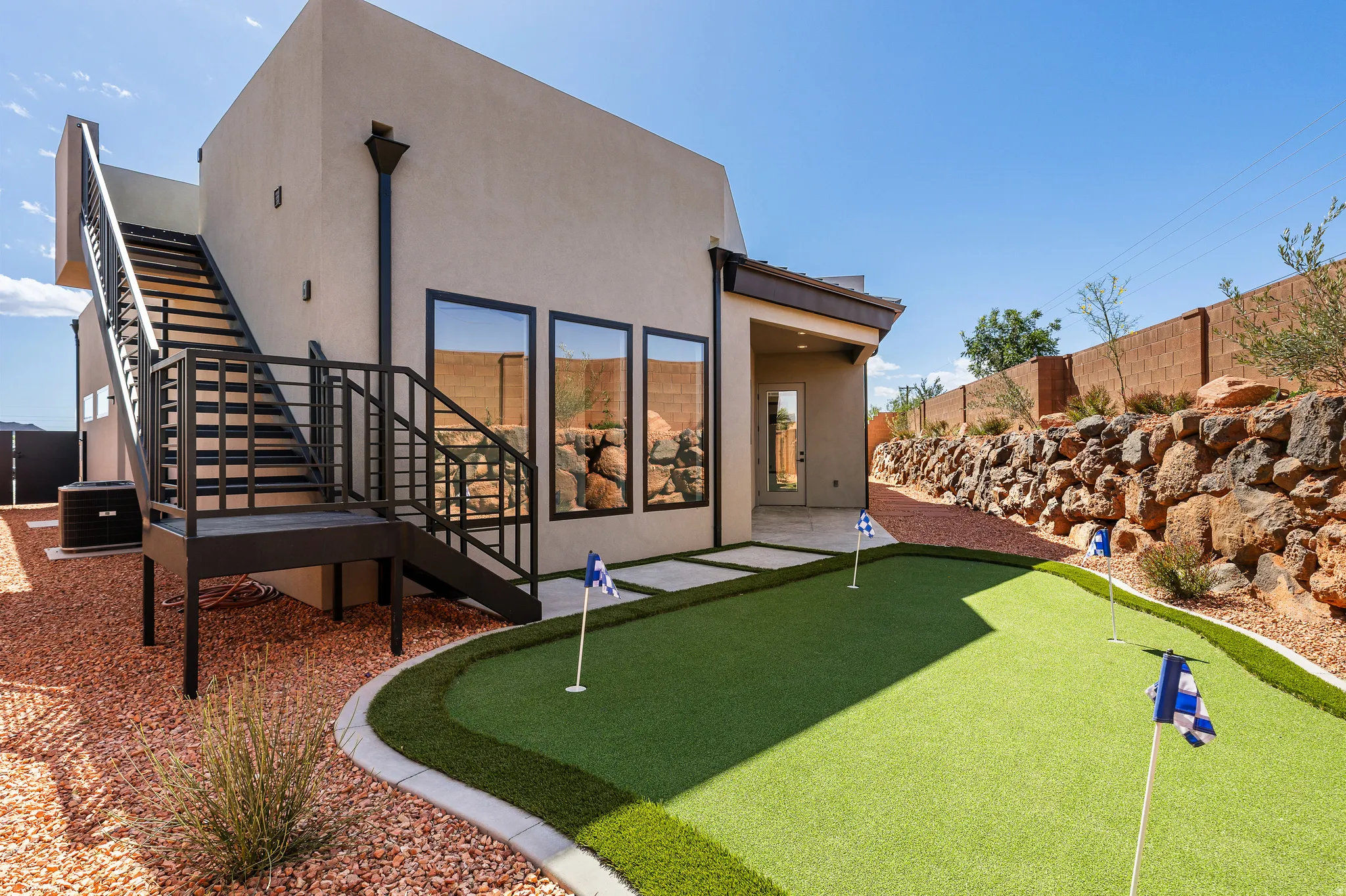 Rear exterior featuring a multi-hole putting green, an external staircase with metal railings, a covered patio area, and a rock retaining wall