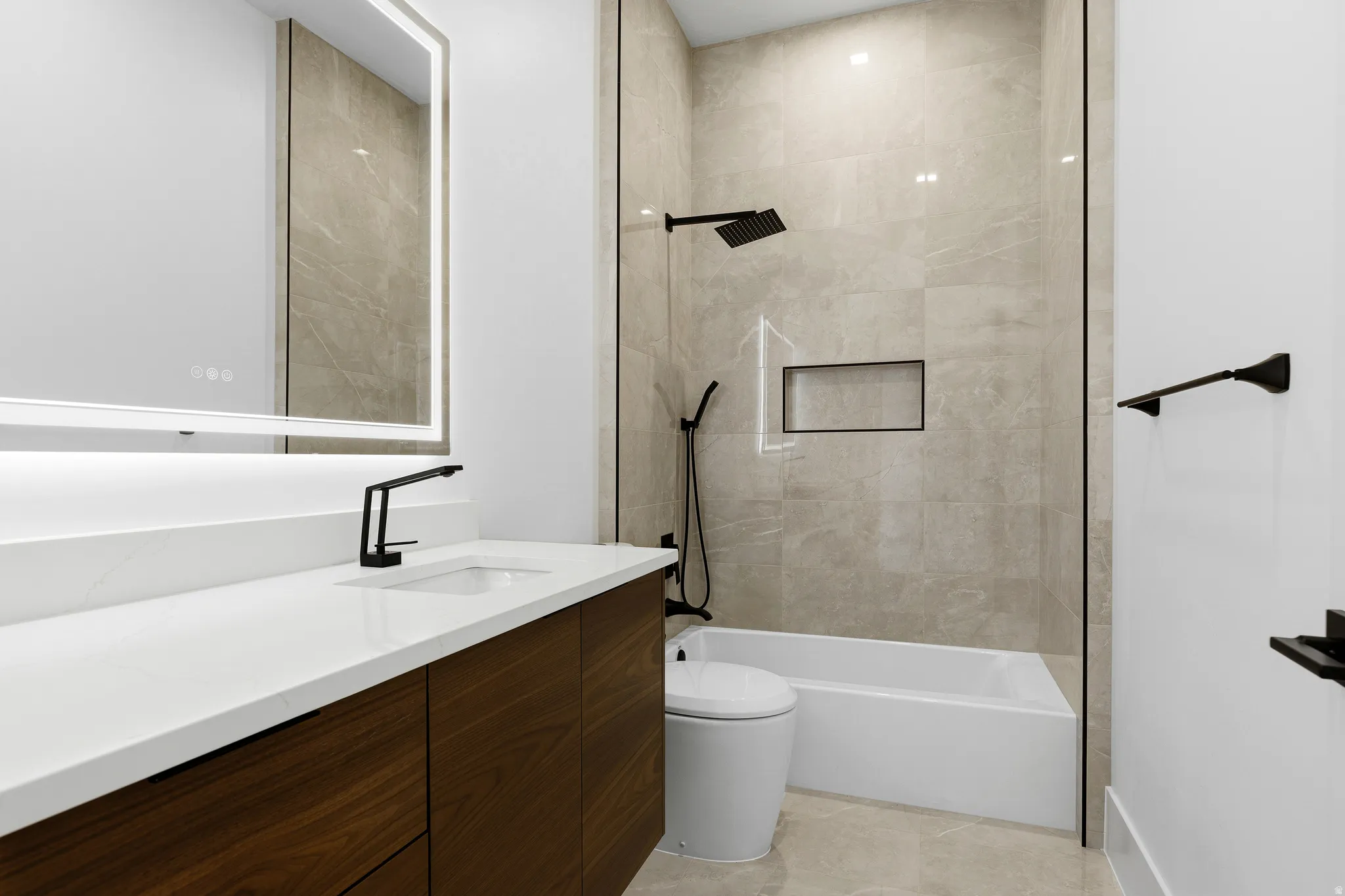 Bathroom featuring a wood-finish vanity with a white countertop and undermount sink, illuminated mirror, matte black fixtures, and a tiled shower with a niche