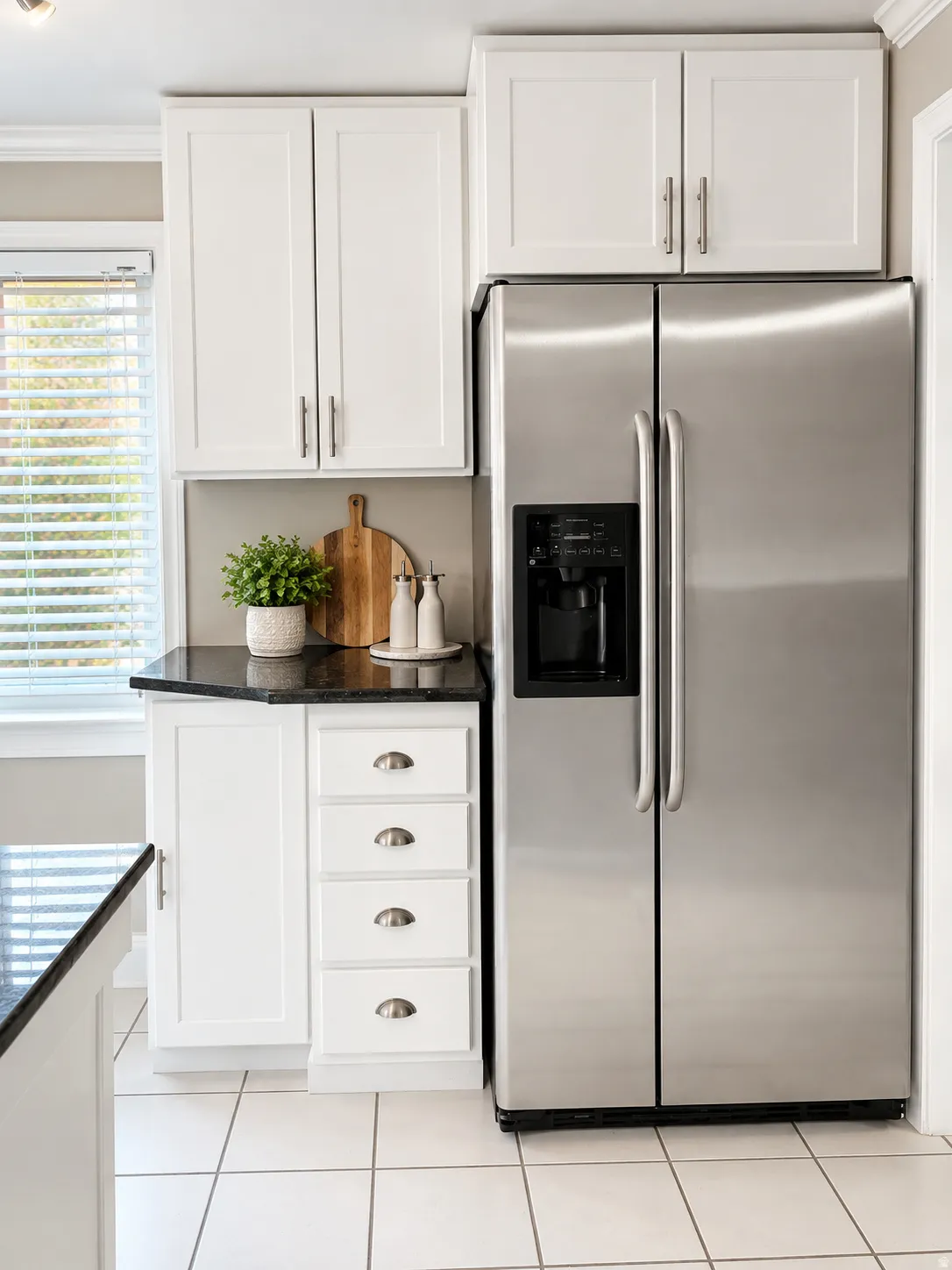 Stainless steel side-by-side refrigerator, white shaker-style cabinetry, dark stone countertop, white tile flooring, and integrated window blinds
