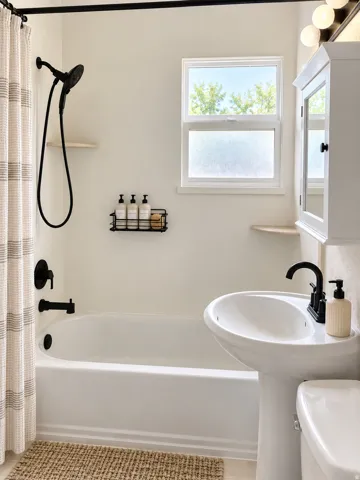 Pedestal sink with a matte black faucet, white bathtub with matte black fixtures, white-framed window, and a white wall-mounted cabinet