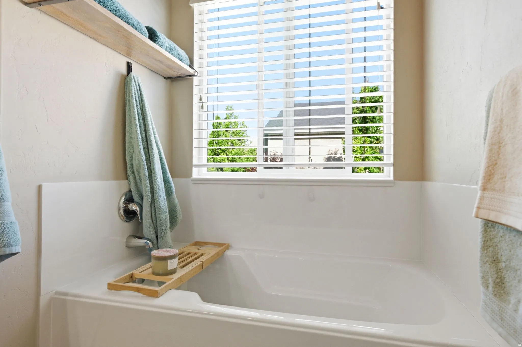 Deep soaking tub featuring chrome fixtures, a window with horizontal blinds, and a built-in wood-finish shelving unit