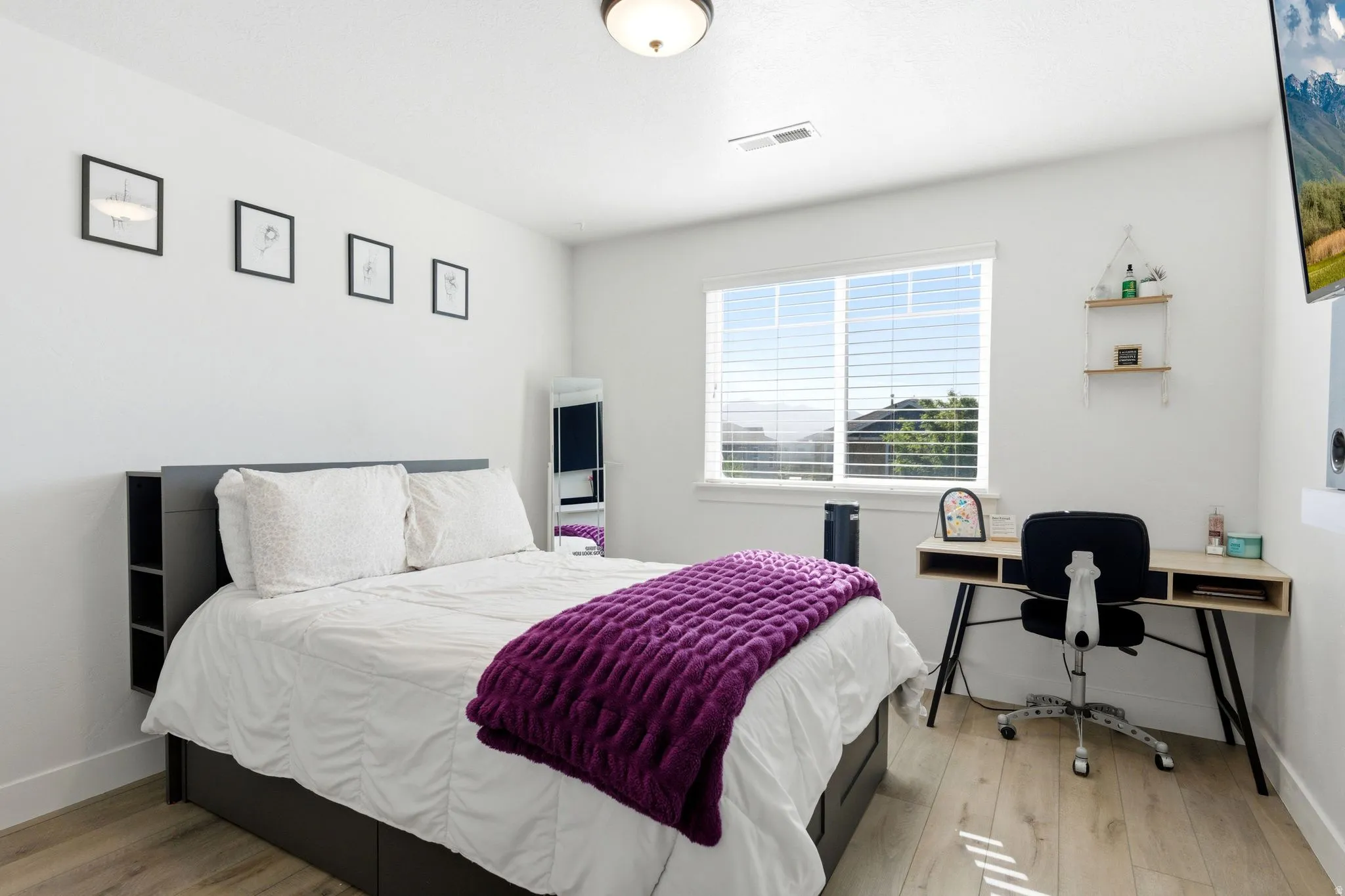 Bright bedroom with light wood-finish flooring, white walls, and a large window with white blinds
