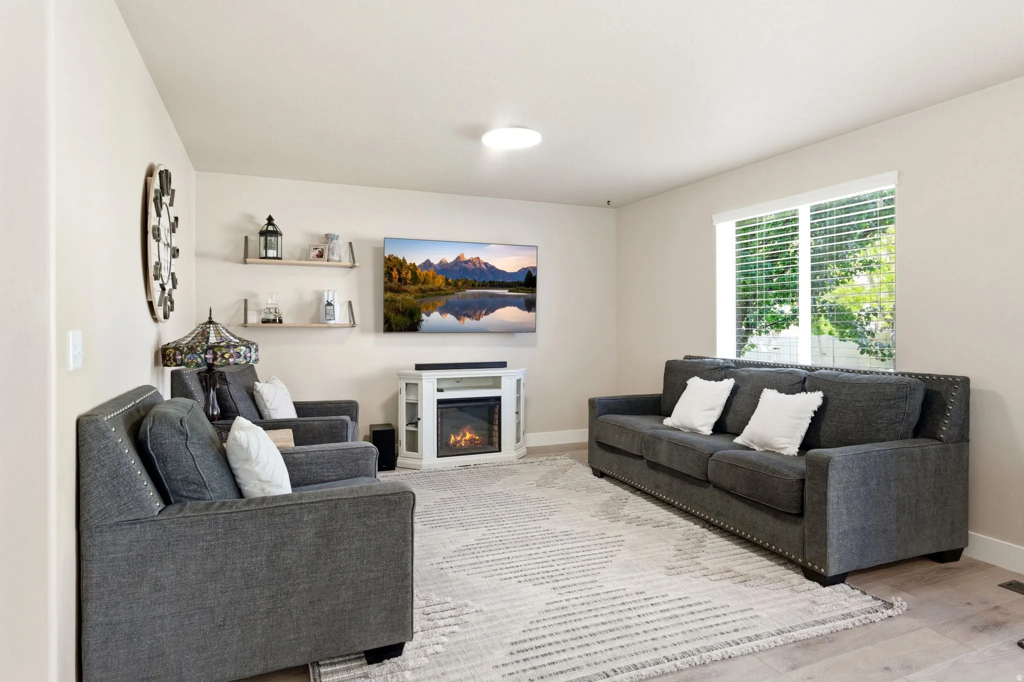 Living area featuring light wood-finish flooring, a contemporary ceiling light fixture, and a large window with horizontal blinds