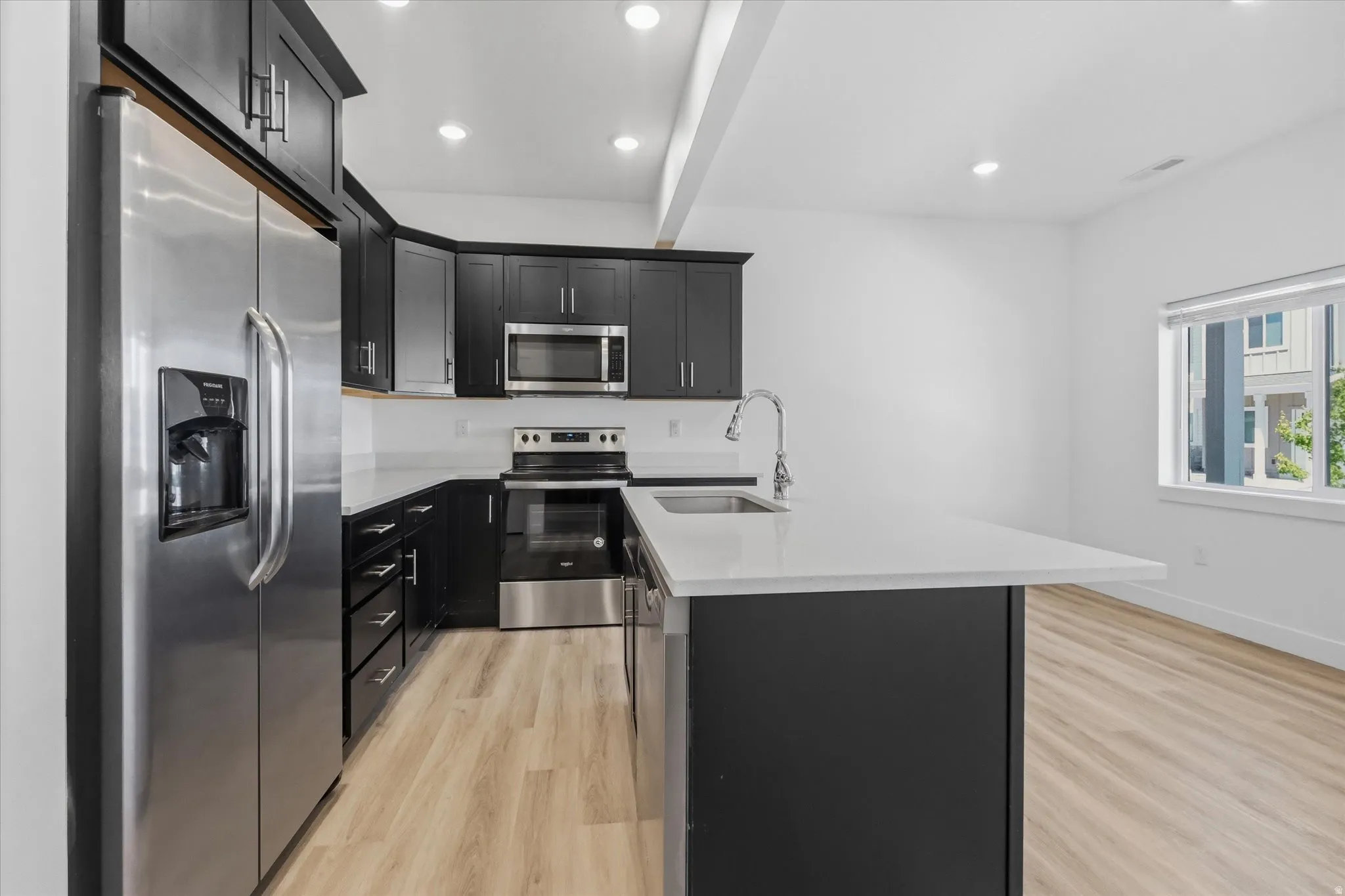 Modern kitchen featuring dark cabinetry, stainless steel appliances, light-toned countertops, and wood-finish flooring