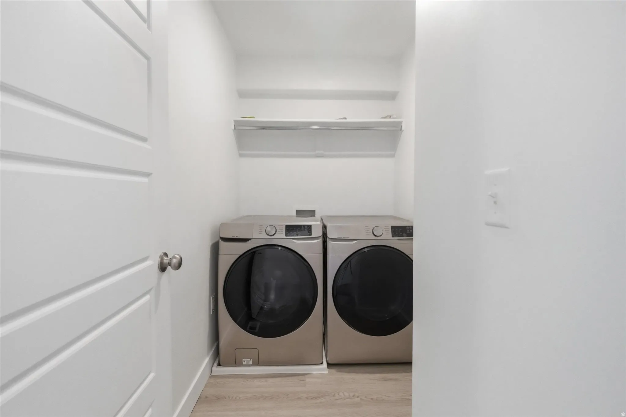 Dedicated laundry area featuring modern front-load appliances, overhead shelving, and wood-finish flooring