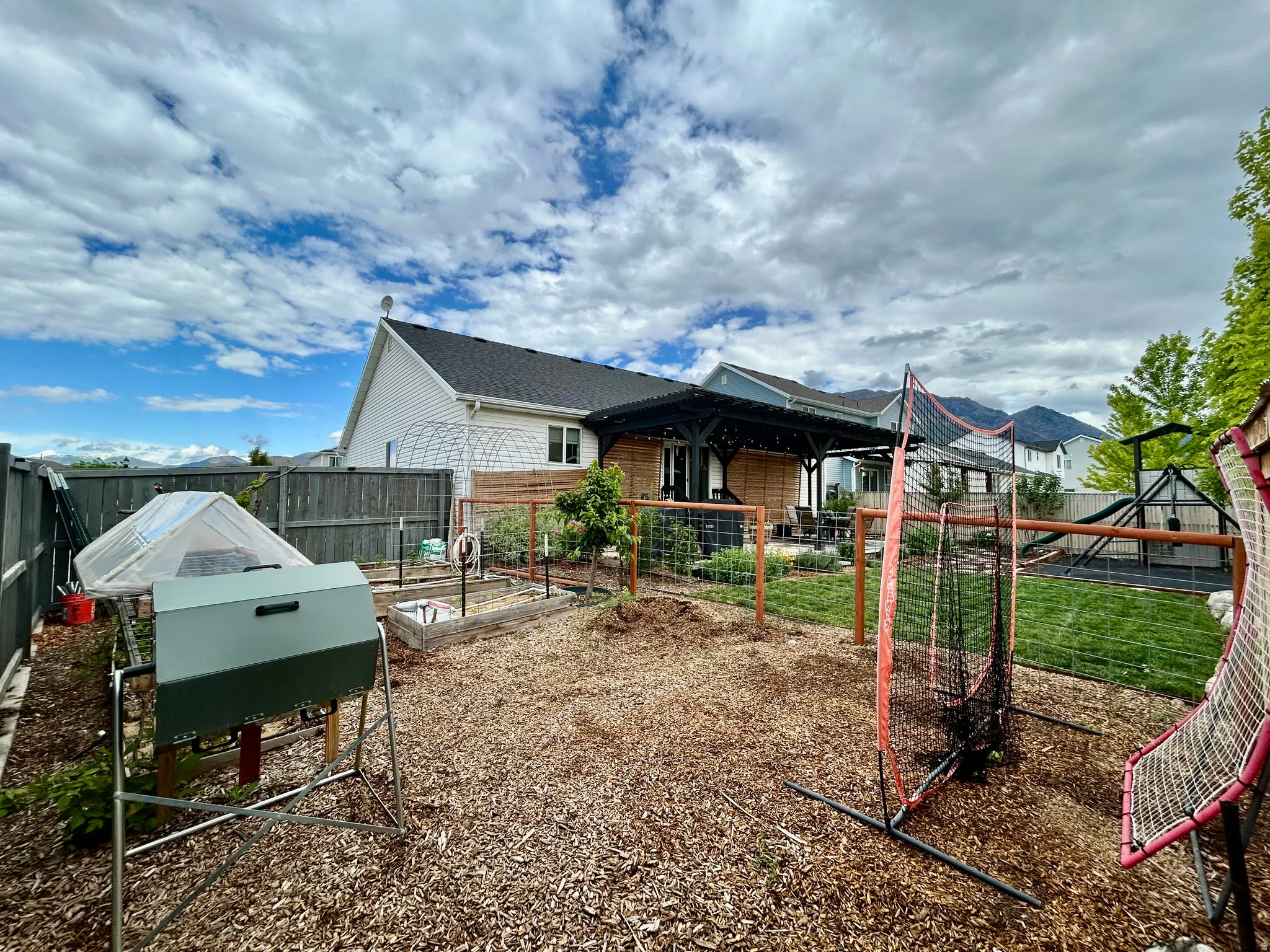 Spacious rear yard featuring an expansive mulched area, multiple raised garden beds, a covered patio with string lighting, and a lawn area with a play structure