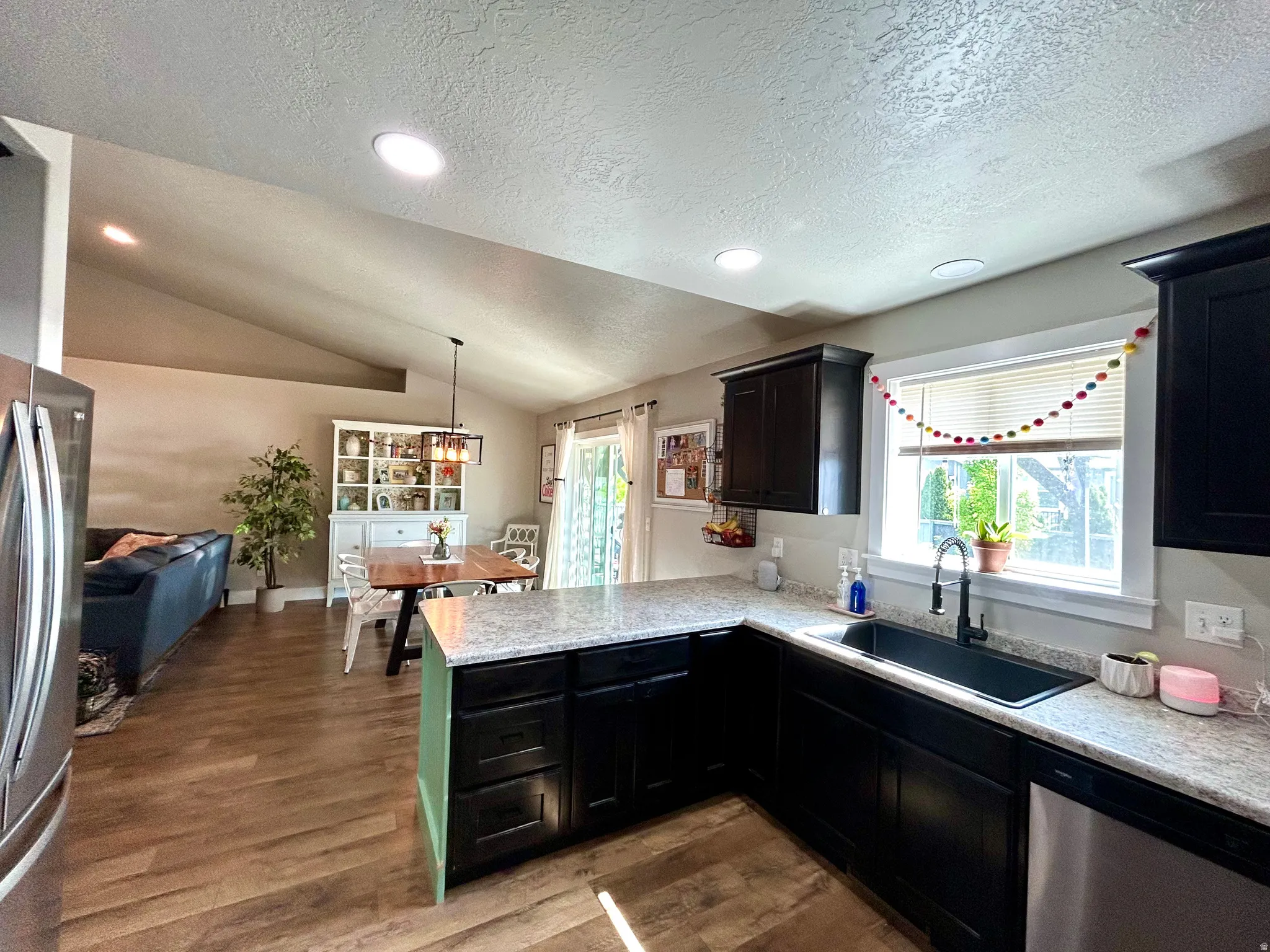 Open-concept kitchen and living area featuring wood-finish flooring, vaulted ceilings with recessed lighting, dark cabinetry, light-toned countertops, and a stainless steel dishwasher