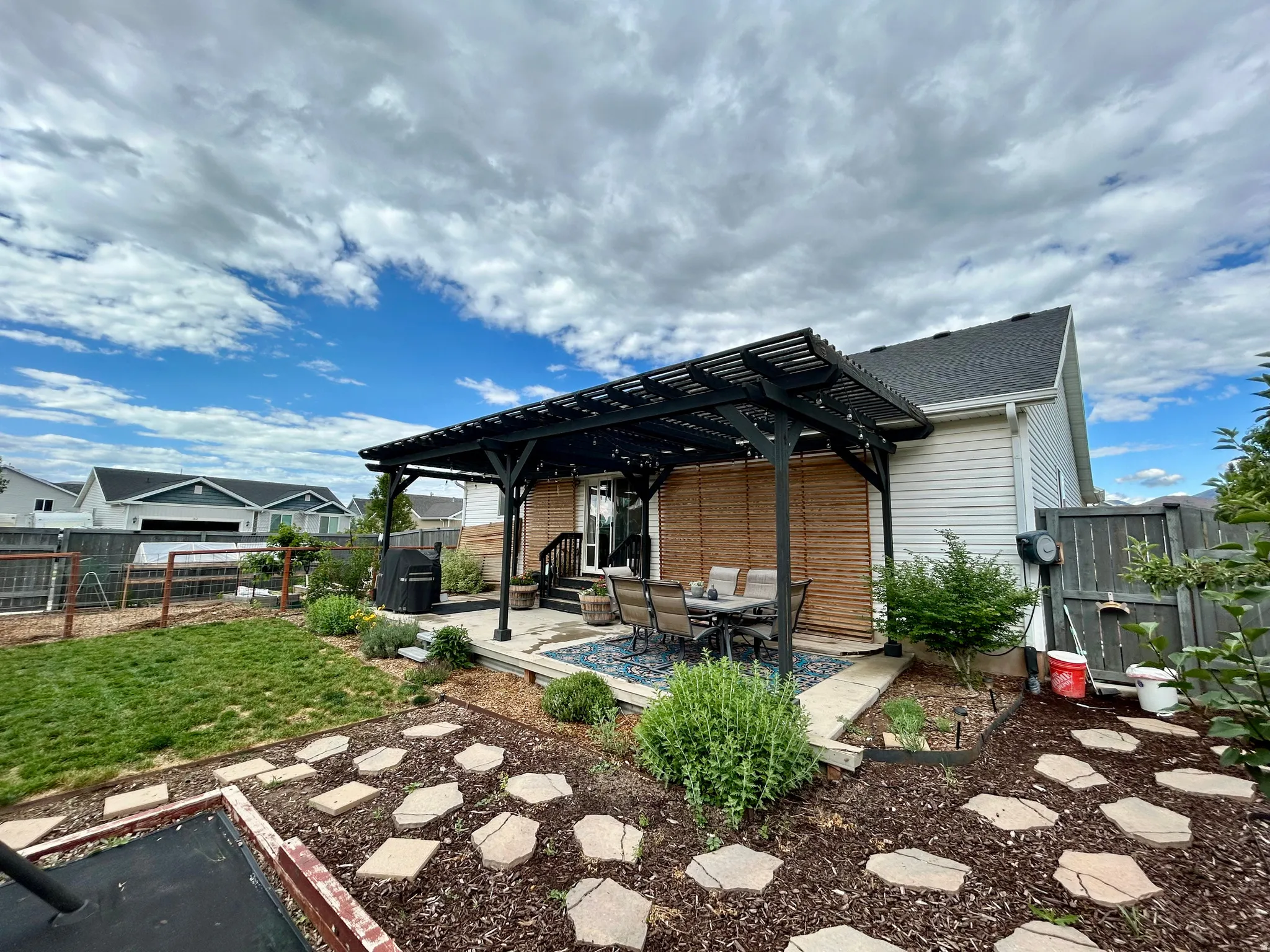 Expansive backyard featuring a dark-toned pergola with slatted roof, a concrete patio area, and a stone paver pathway