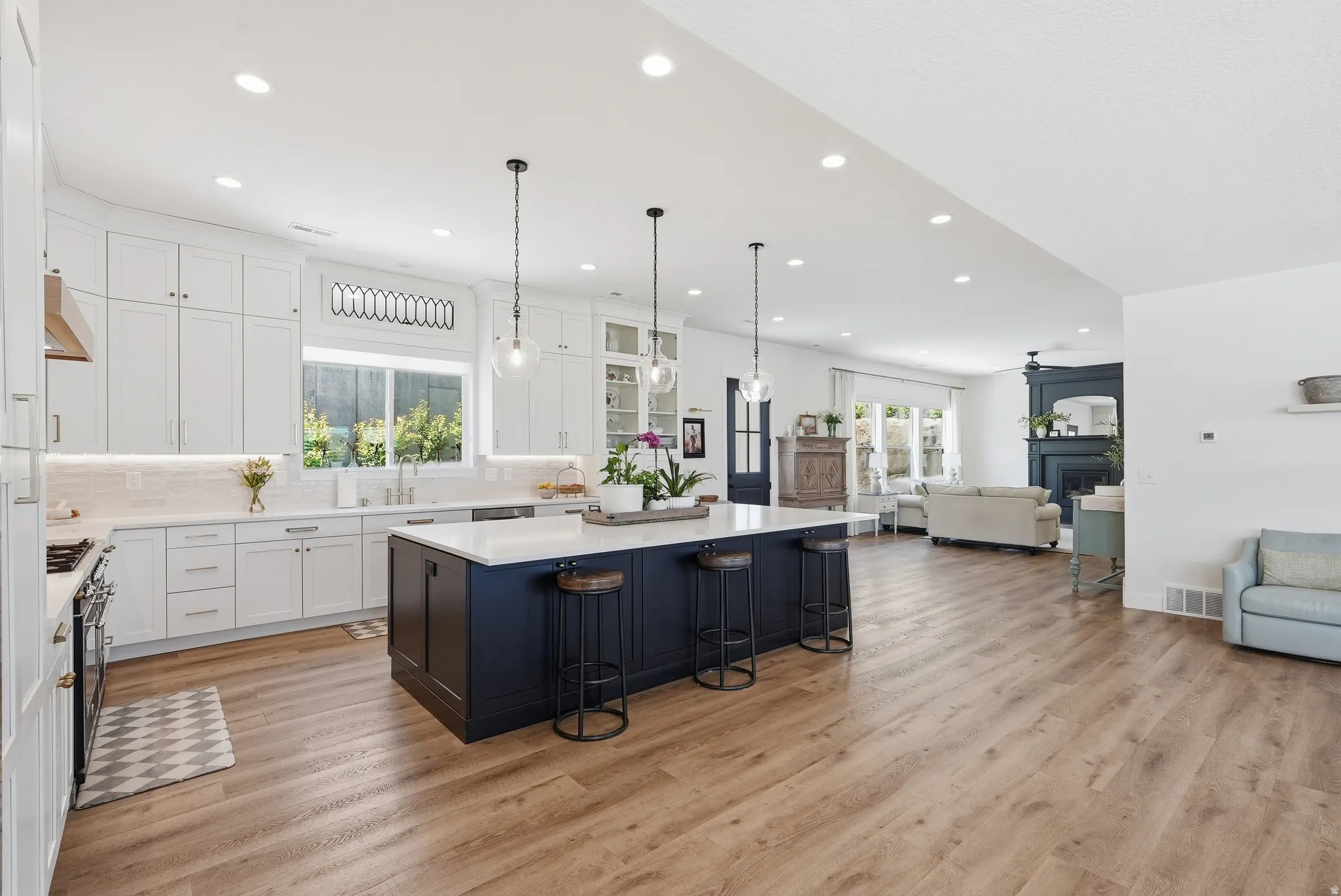 Spacious kitchen featuring an expansive island with seating, white shaker cabinetry, quartz countertops, and wood-finish flooring extending into the living area with a dark-toned fireplace surround