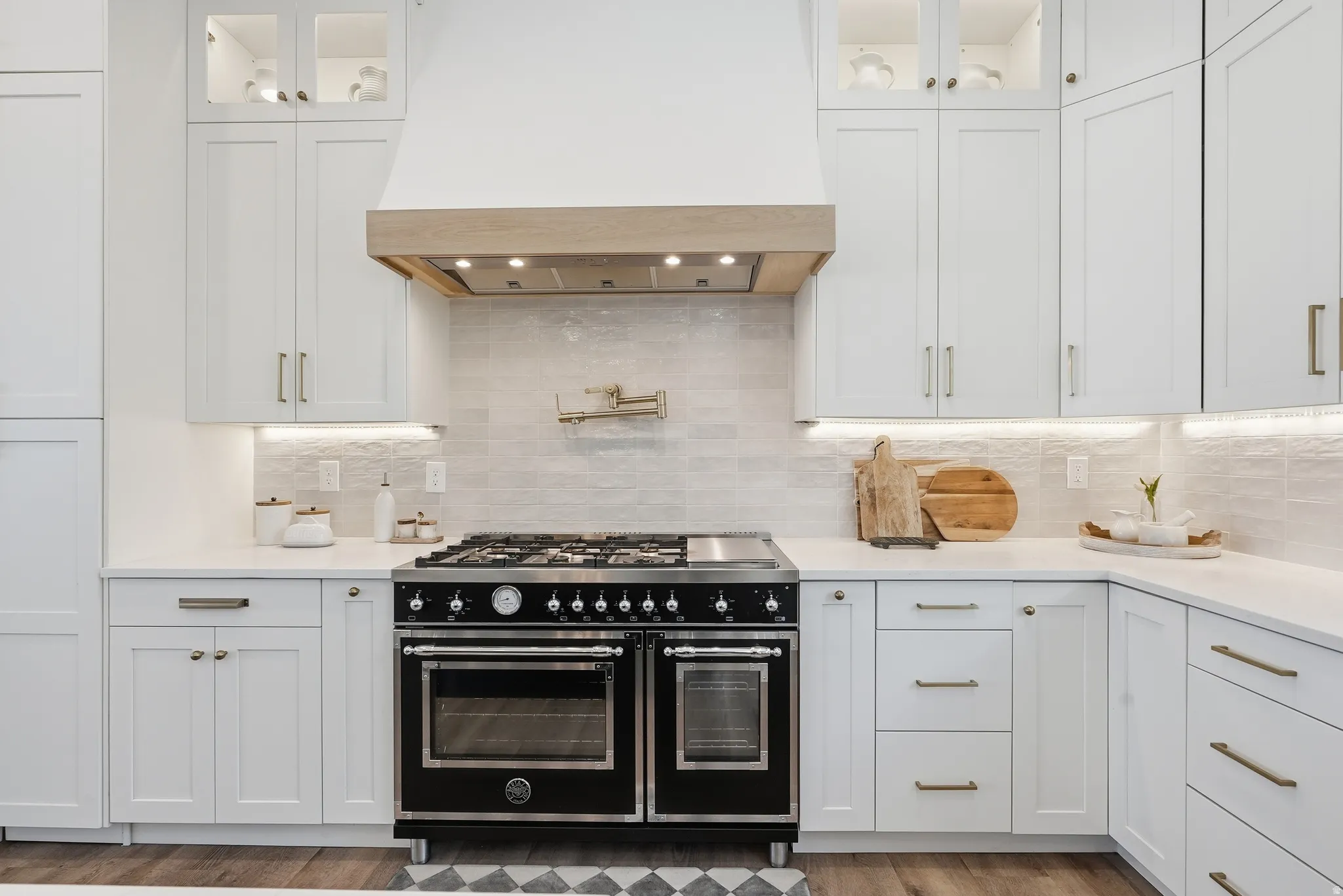 Gourmet kitchen featuring a professional-grade black range, custom wood-paneled range hood, white shaker cabinetry with gold-tone hardware, textured tile backsplash, and under-cabinet lighting