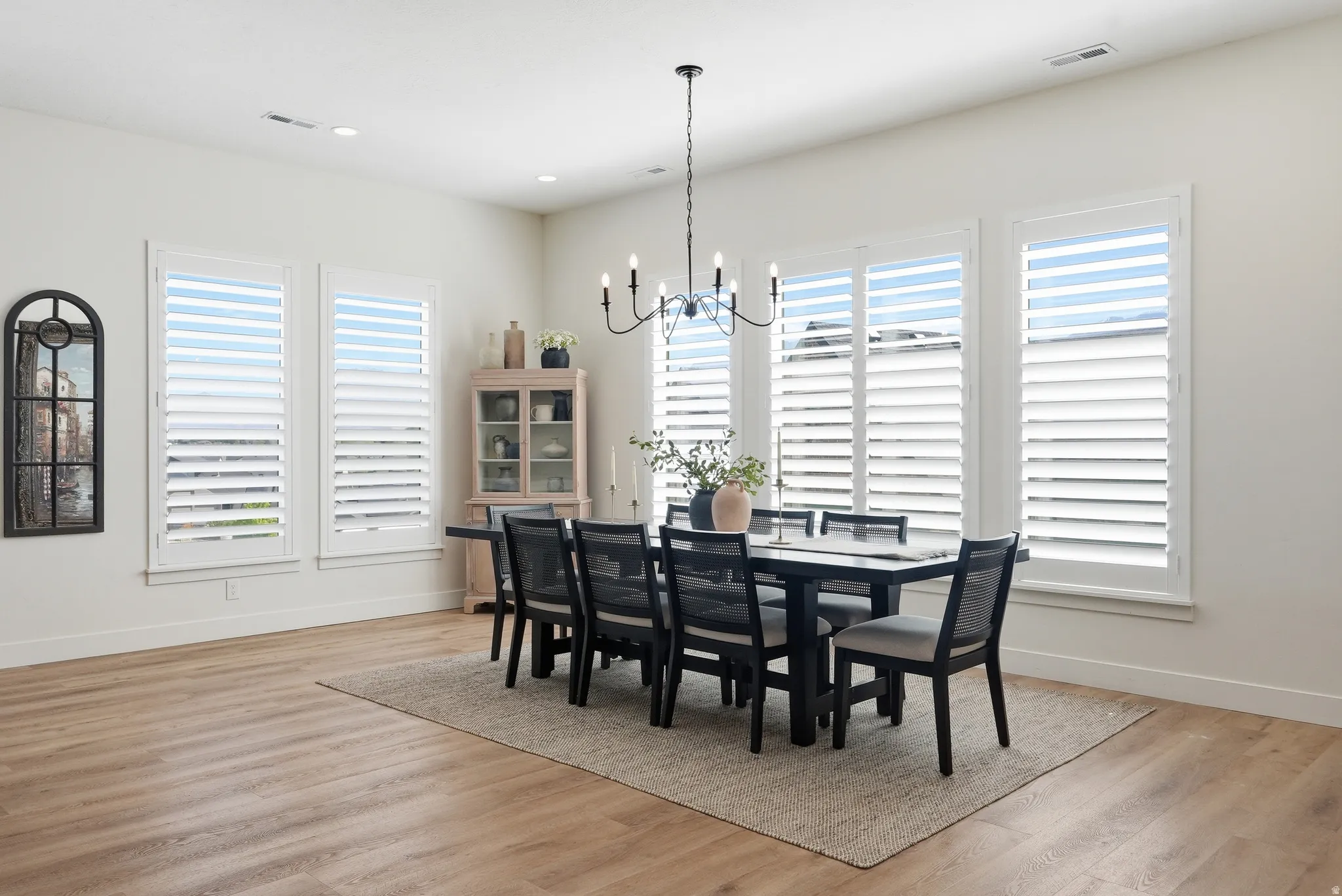 Spacious dining area featuring large windows with plantation shutters, LVP flooring, a contemporary chandelier, and a neutral wall palette
