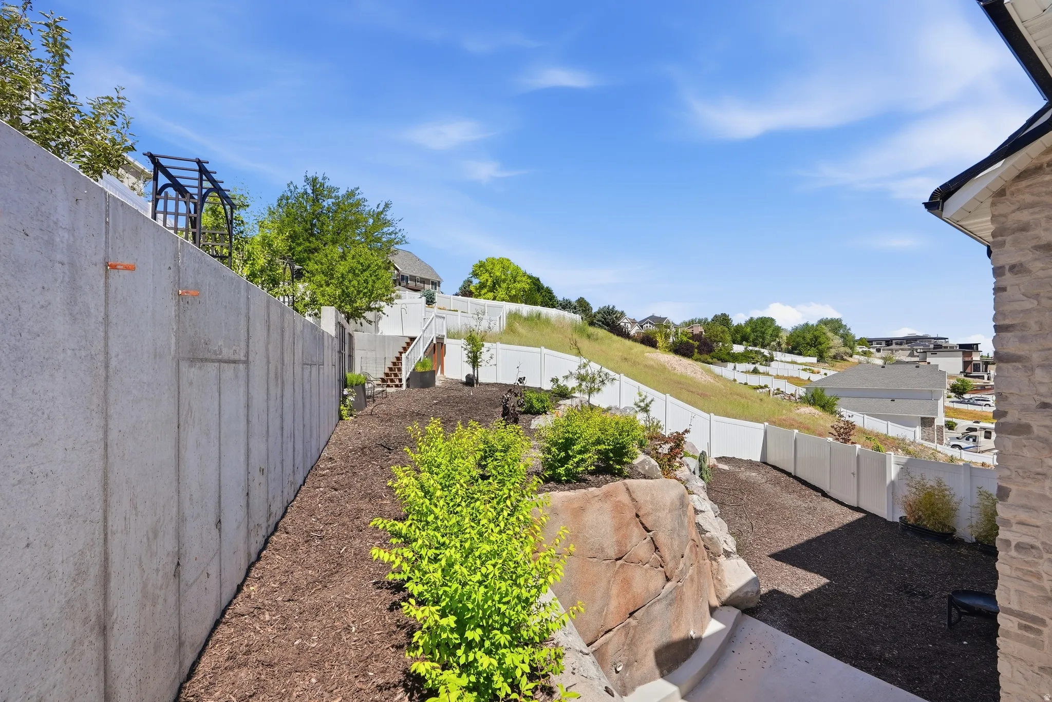 Terraced hillside landscaping featuring a concrete retaining wall, a multi-level white privacy fence, an integrated rock feature, and planted shrubs with bark mulch