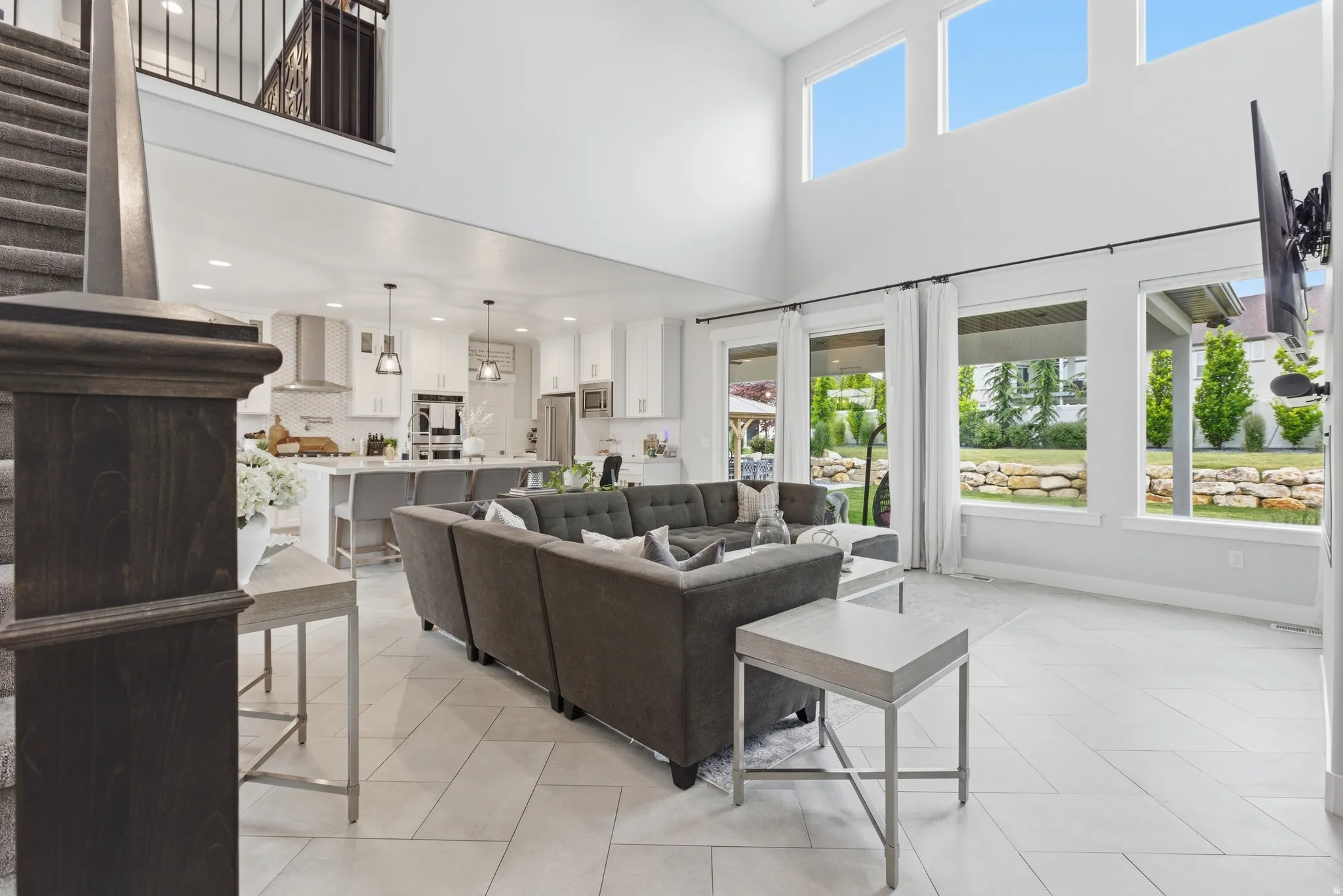 Vaulted ceiling great room featuring expansive windows, light tile flooring, an open-concept kitchen with a large island, and a carpeted staircase with dark wood newel posts