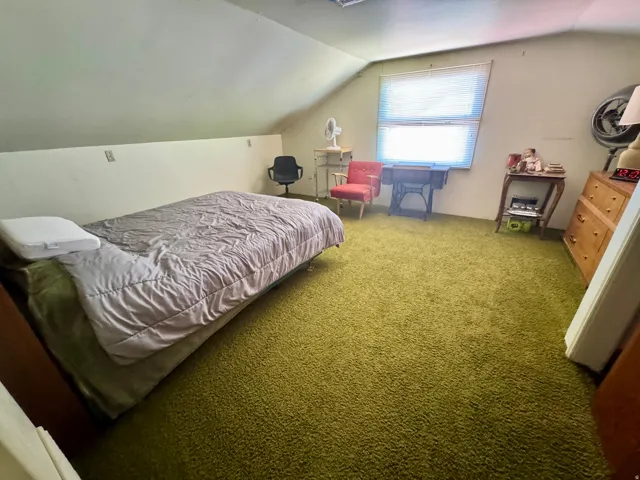 Room with textured green carpeting, sloped ceilings, and a window with blinds