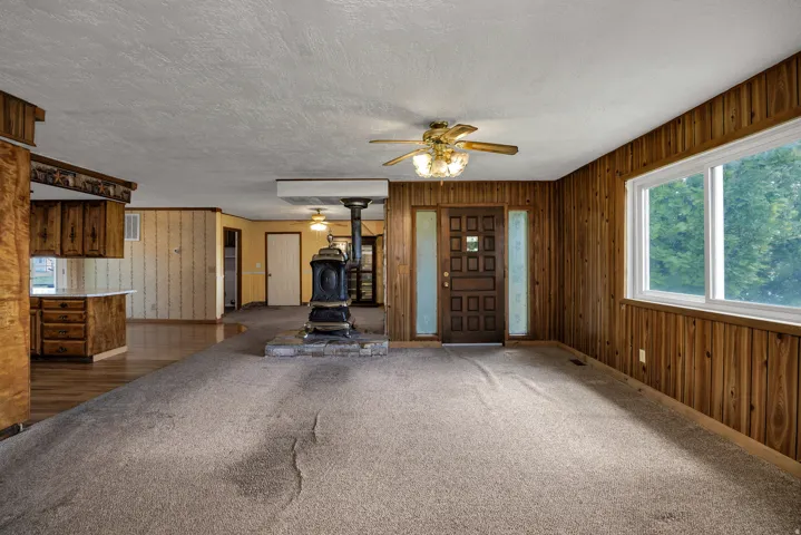 Living area featuring a freestanding stove on a stone hearth, wood-paneled walls, a large window, and a ceiling fan light fixture