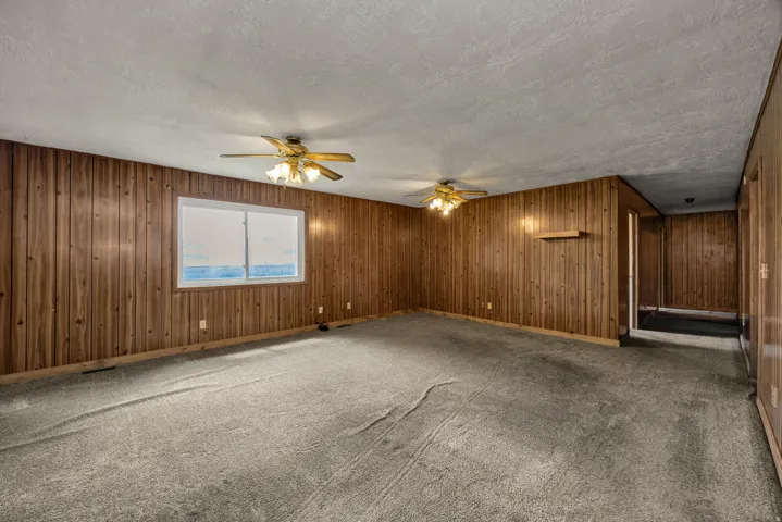 Spacious room featuring wood paneling, a large window, two ceiling fans with integrated lighting, and carpeted flooring