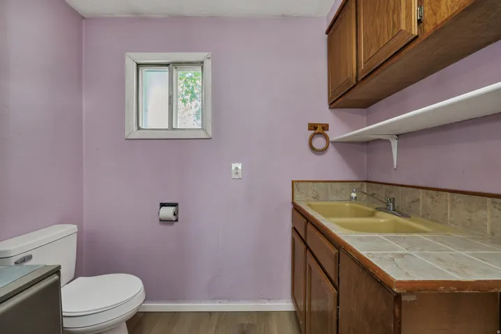 Utility space featuring wood-finish flooring, a white window frame, a white toilet, brown lower cabinetry with a tiled countertop, and an upper cabinet with an open shelf