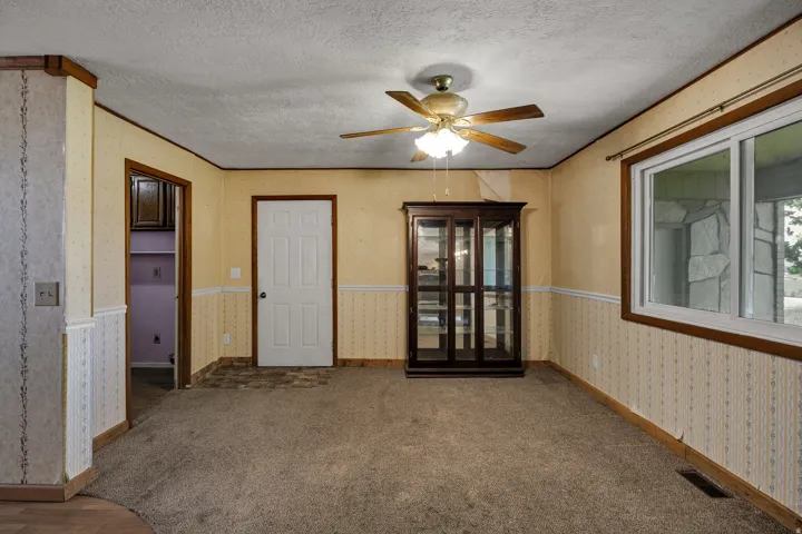 Carpeted interior space featuring a ceiling fan with integrated lighting, white-framed window with wood trim, and a white six-panel door