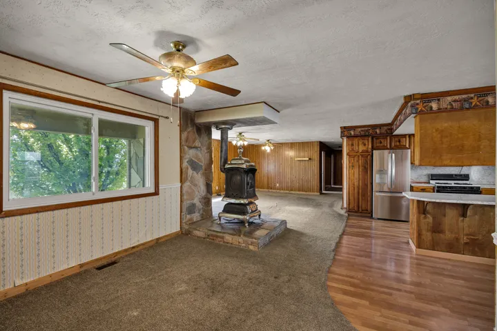Living area featuring a freestanding wood-burning stove on a stone hearth, with a large window, carpet flooring, and a ceiling fan