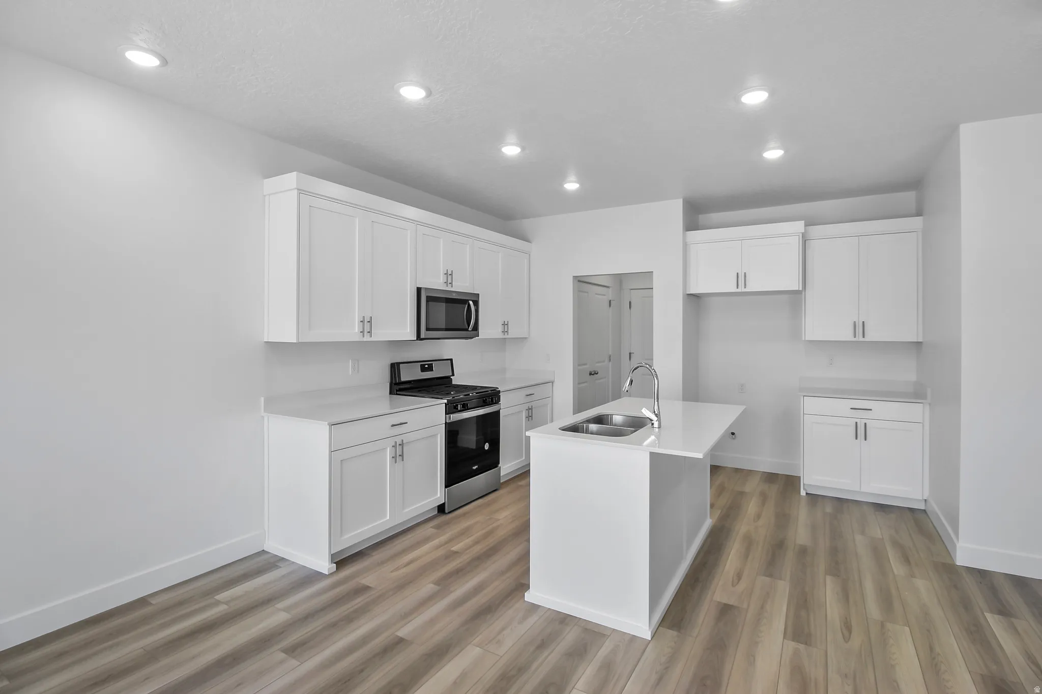 Kitchen featuring white shaker cabinetry, stainless steel appliances, light-toned countertops, a central island with a double basin sink, and wood-finish flooring