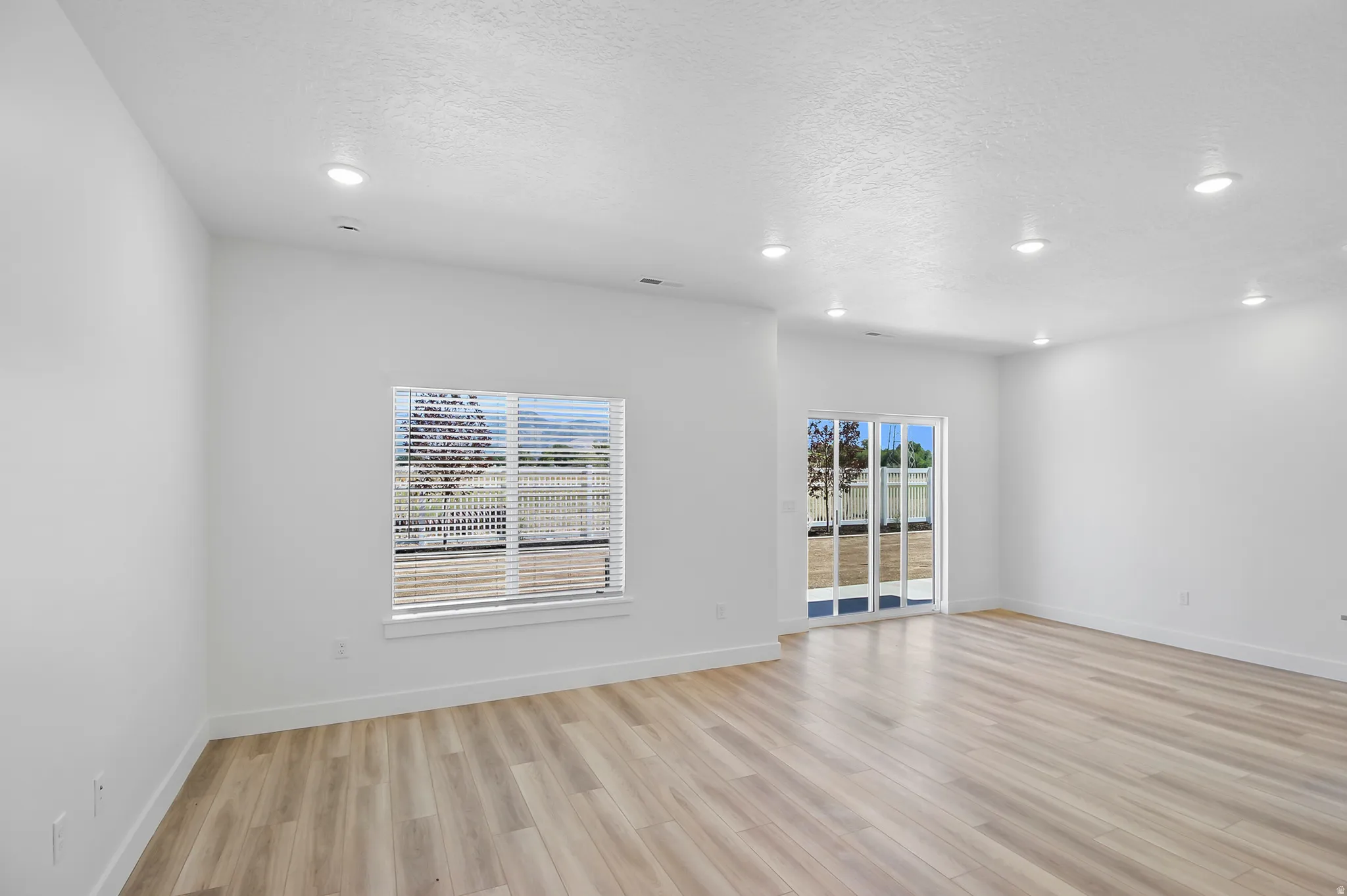 Spacious room featuring light wood-finish flooring, recessed ceiling lighting, a large window with blinds, and sliding glass doors providing access to the outdoors