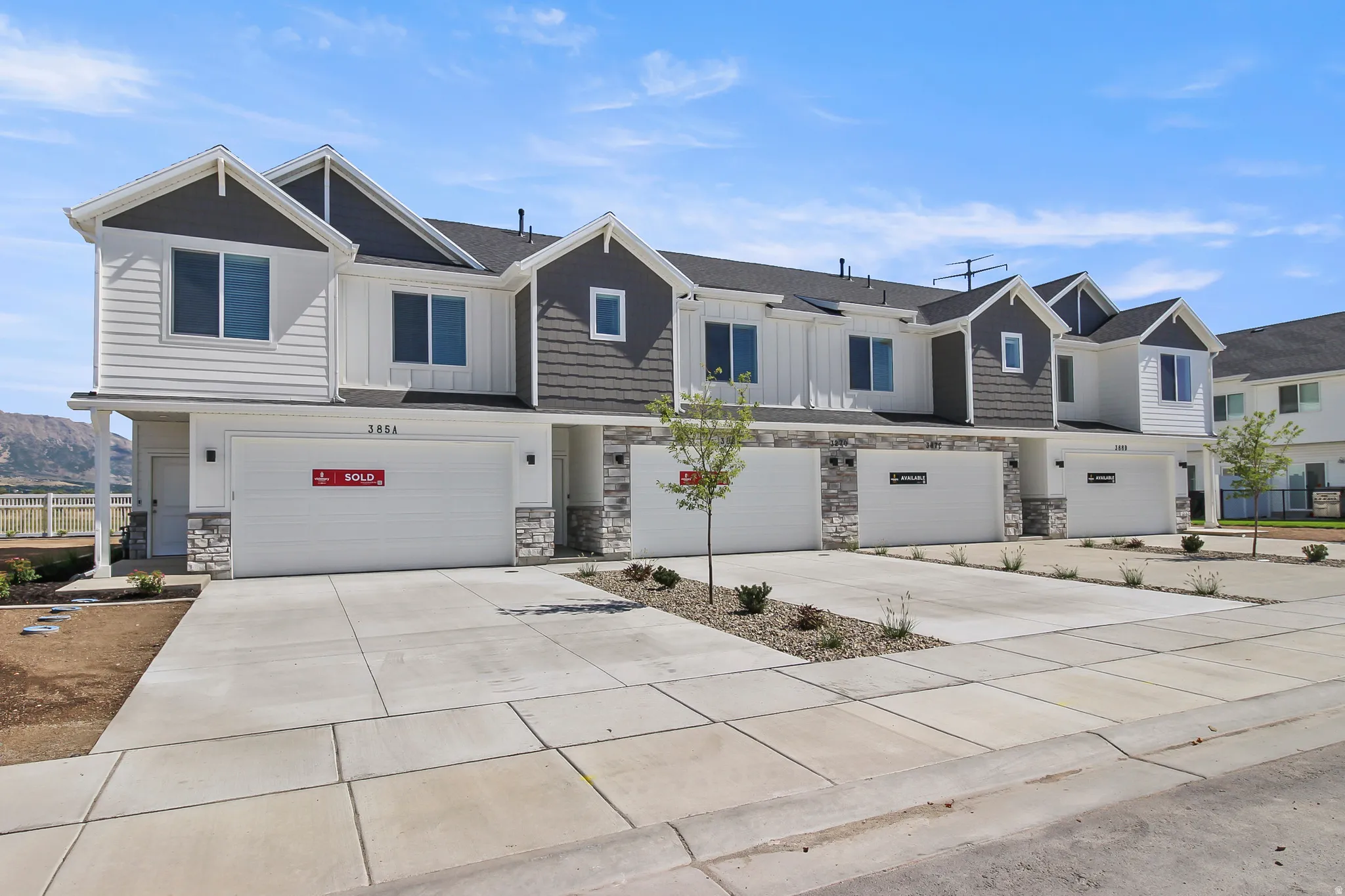 Contemporary townhome-style residences featuring white vertical and horizontal siding accents, dark shingle roofs, stone veneer facades, concrete driveways, and attached garages