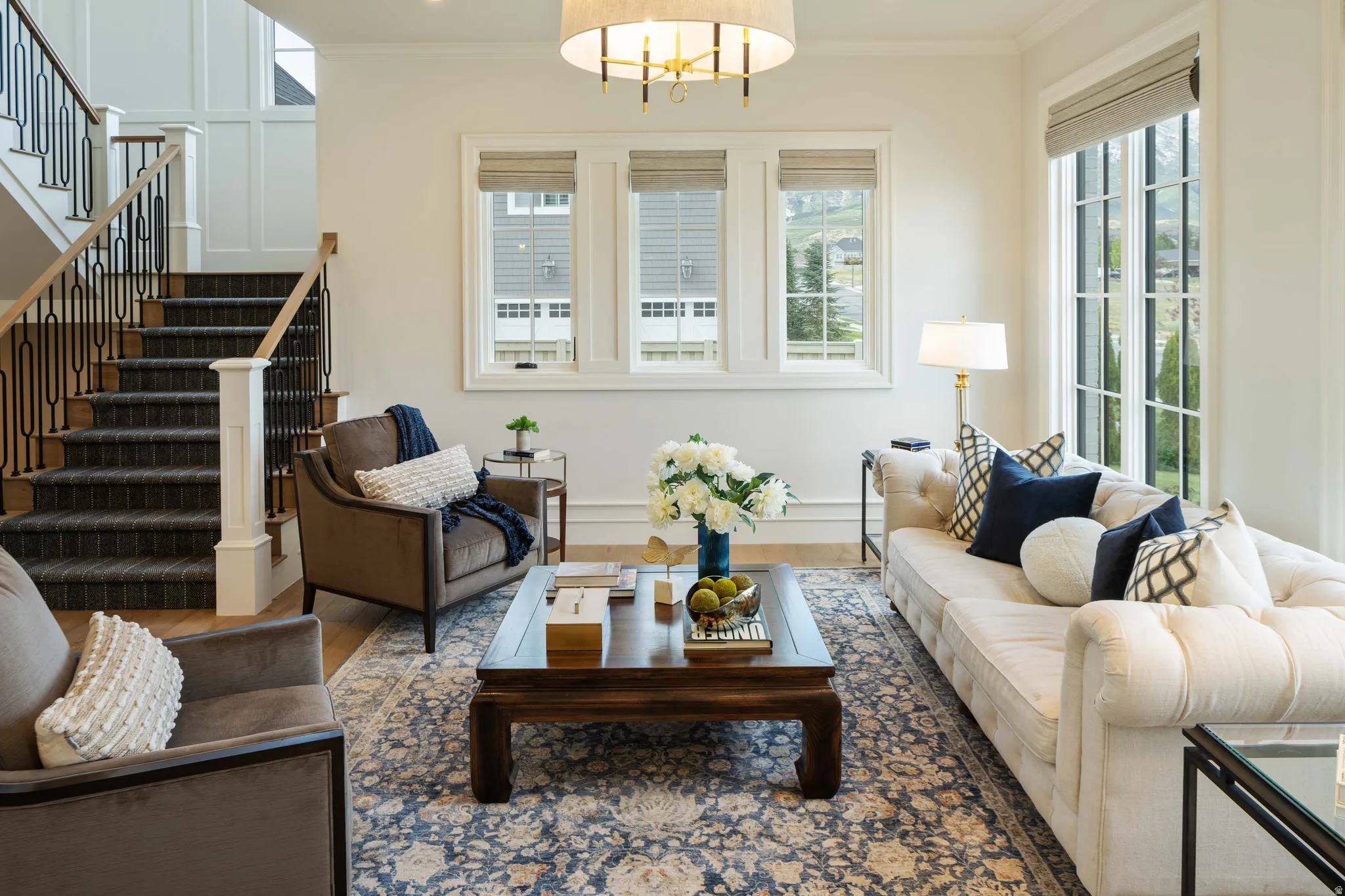 Open living area featuring light wood-finish flooring, a grand staircase with dark metal balusters and wood handrails, large windows with white trim, a decorative chandelier, and extensive wall paneling