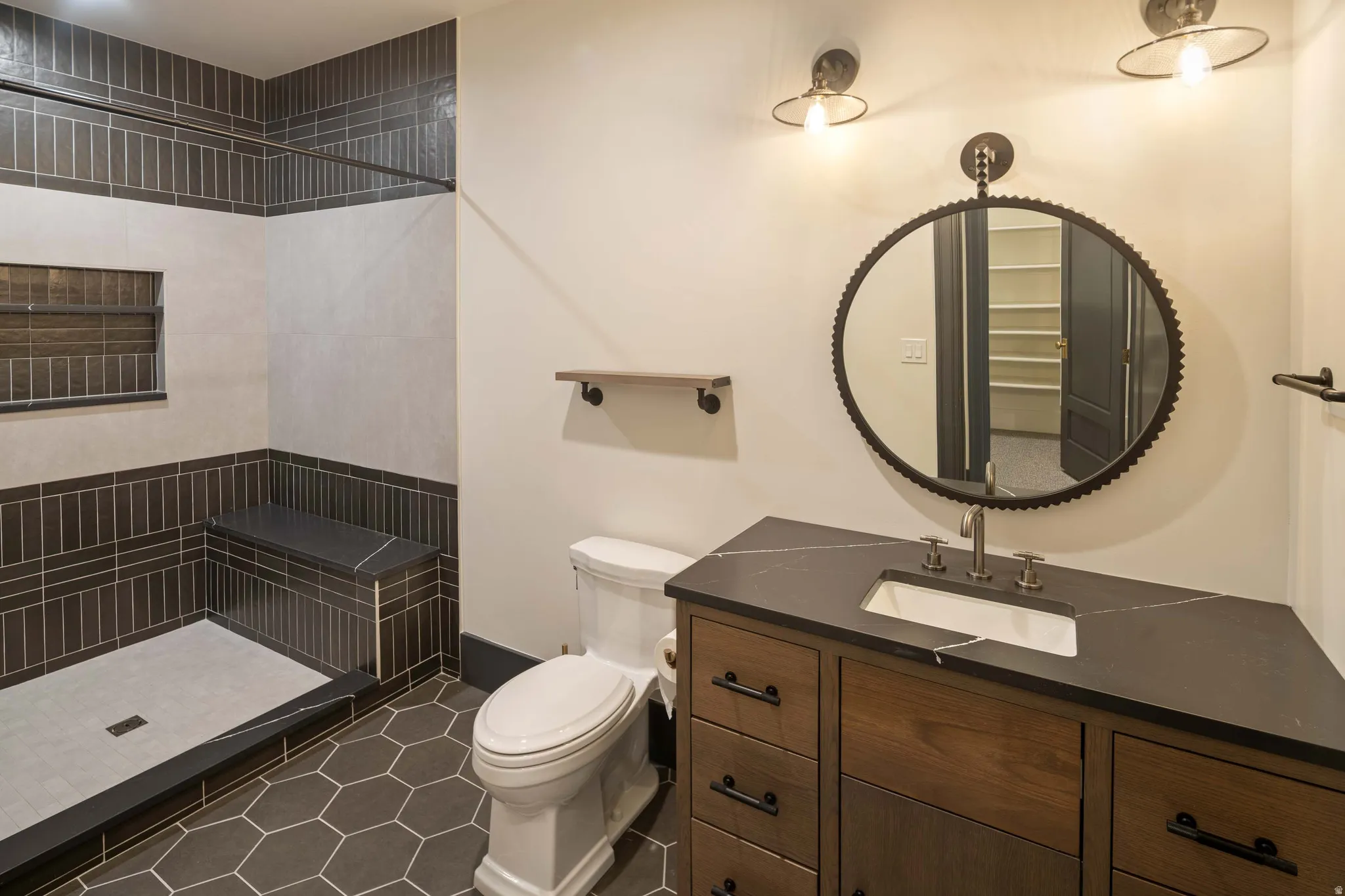 Bathroom featuring hexagonal floor tiling, a walk-in shower with built-in bench and recessed niche, a wood-finish vanity with an integrated sink, and an accent mirror