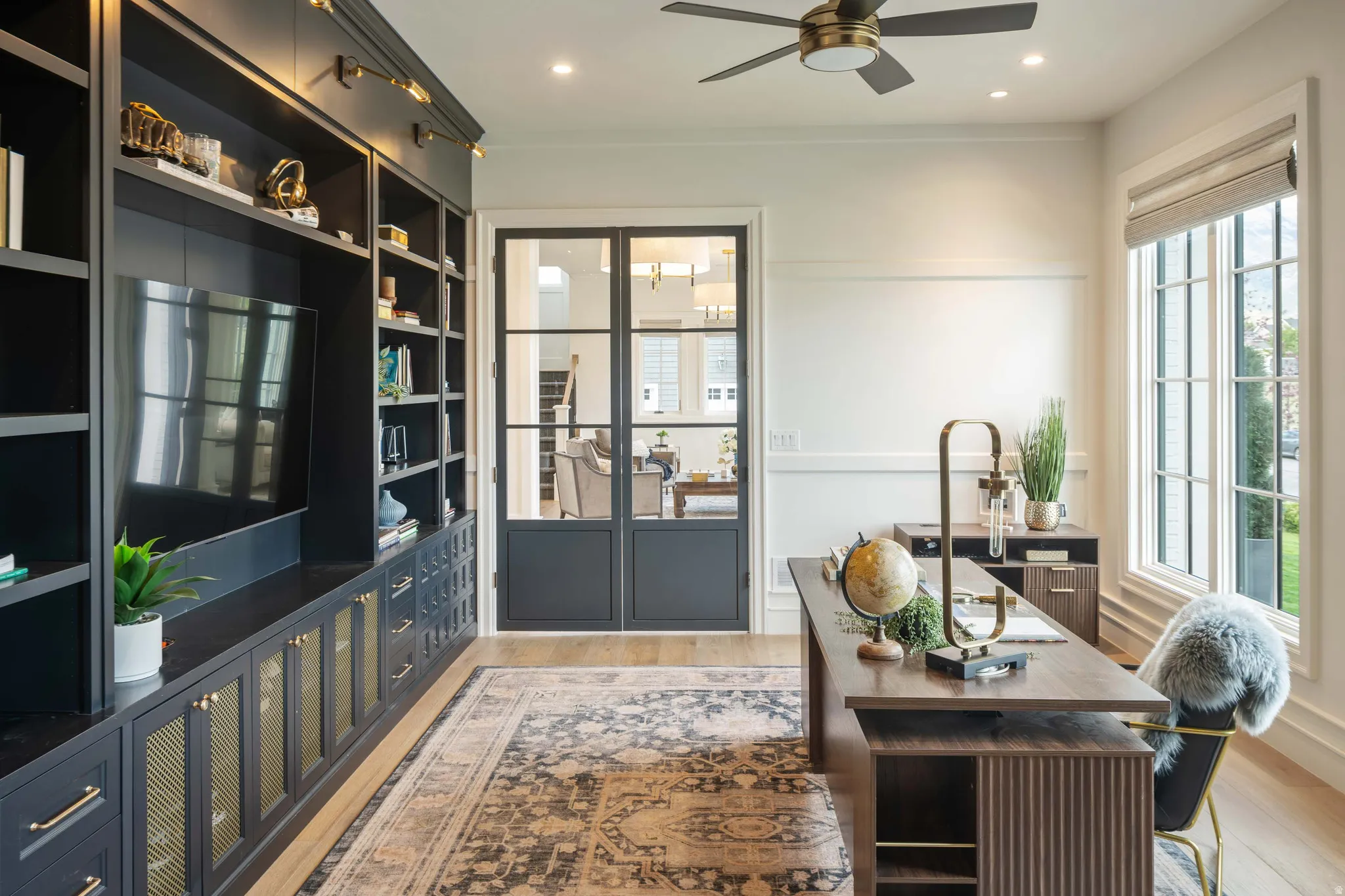 Dedicated office space featuring built-in cabinetry with gold-toned hardware, large multi-pane windows, wood-finish flooring, and a ceiling fan