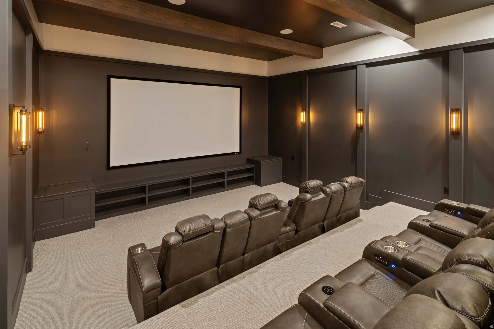 Dedicated media room featuring a large projection screen, built-in shelving, sconce lighting, coffered ceiling with exposed wood beams, and neutral carpeting