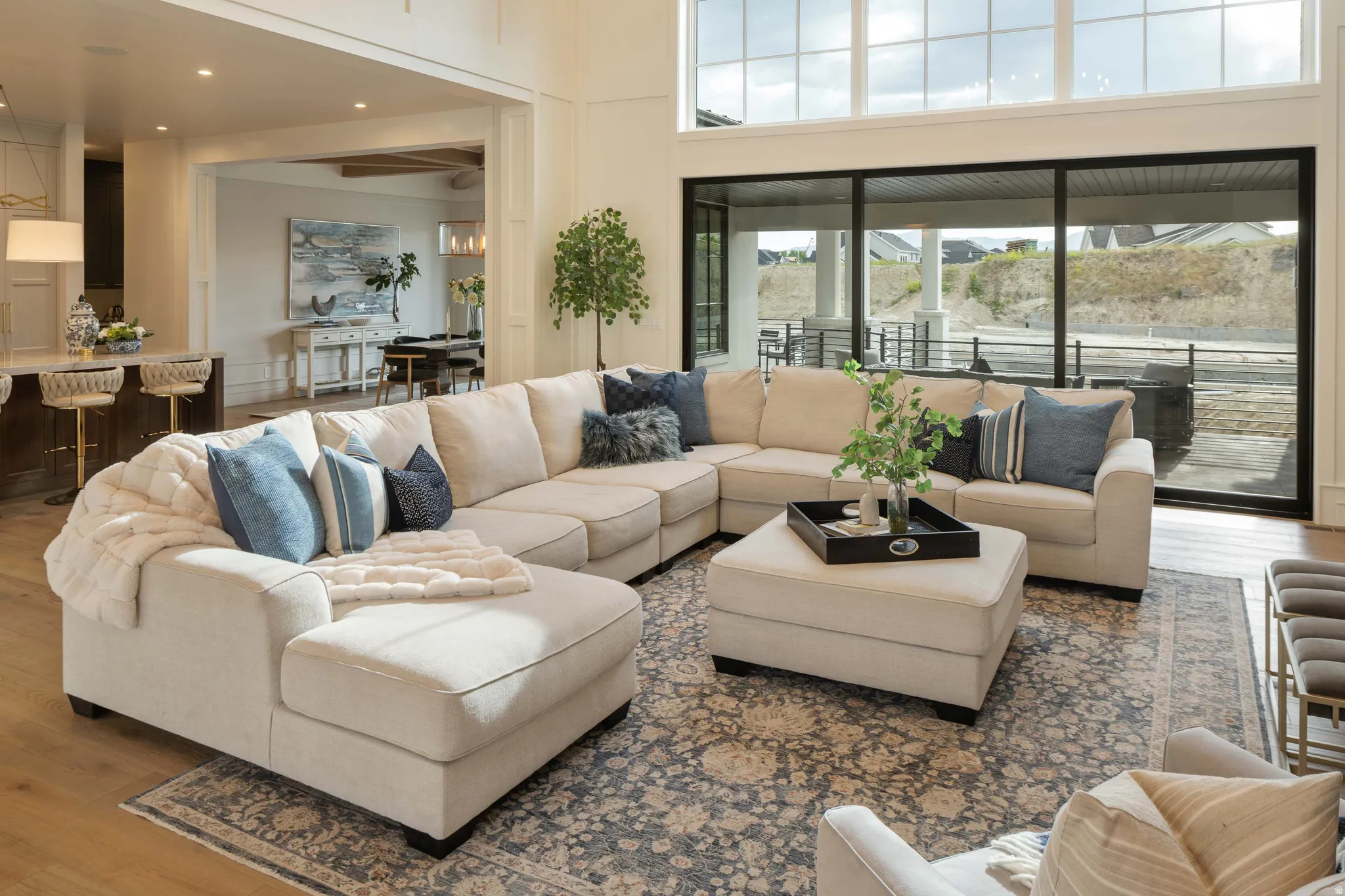 Spacious living area featuring high ceilings, large-format windows with dark frames, wood-finish flooring, and an open concept layout connecting to the kitchen and dining areas