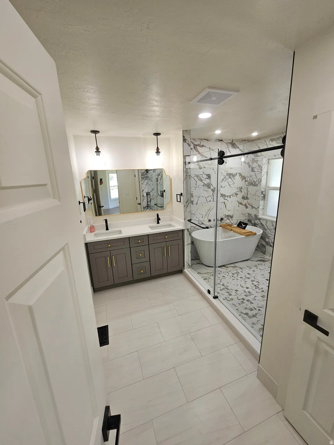 Modern bathroom featuring a dual vanity with gray cabinetry, a freestanding soaking tub, a walk-in shower with a glass enclosure, marble-look tile, and large format floor tiling