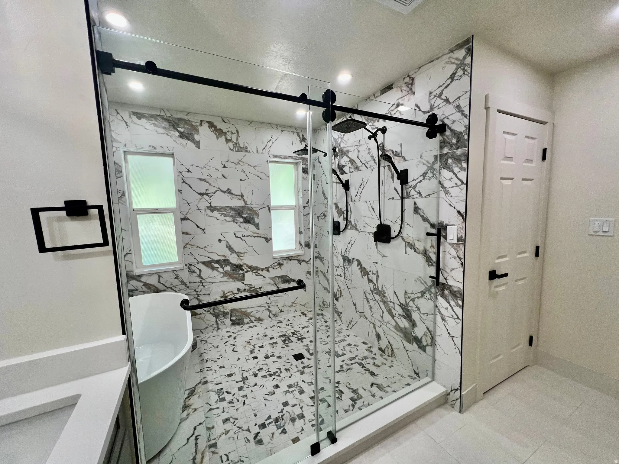 Spacious bathroom featuring a frameless glass shower enclosure with matte black hardware, a freestanding soaking tub, and floor-to-ceiling marble-look tile