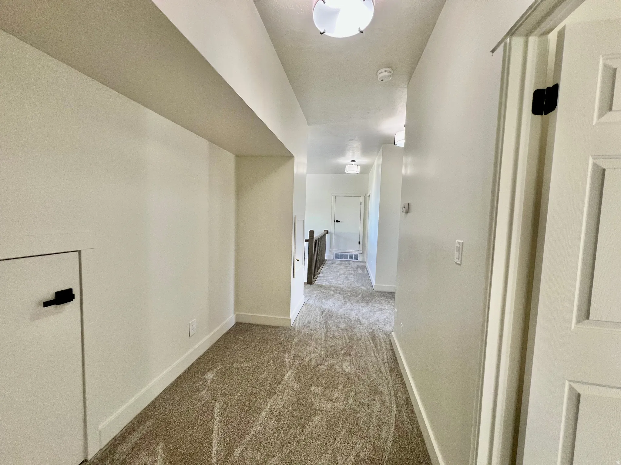 Carpeted hallway featuring bright overhead lighting, white walls, and a built-in access panel with a black latch