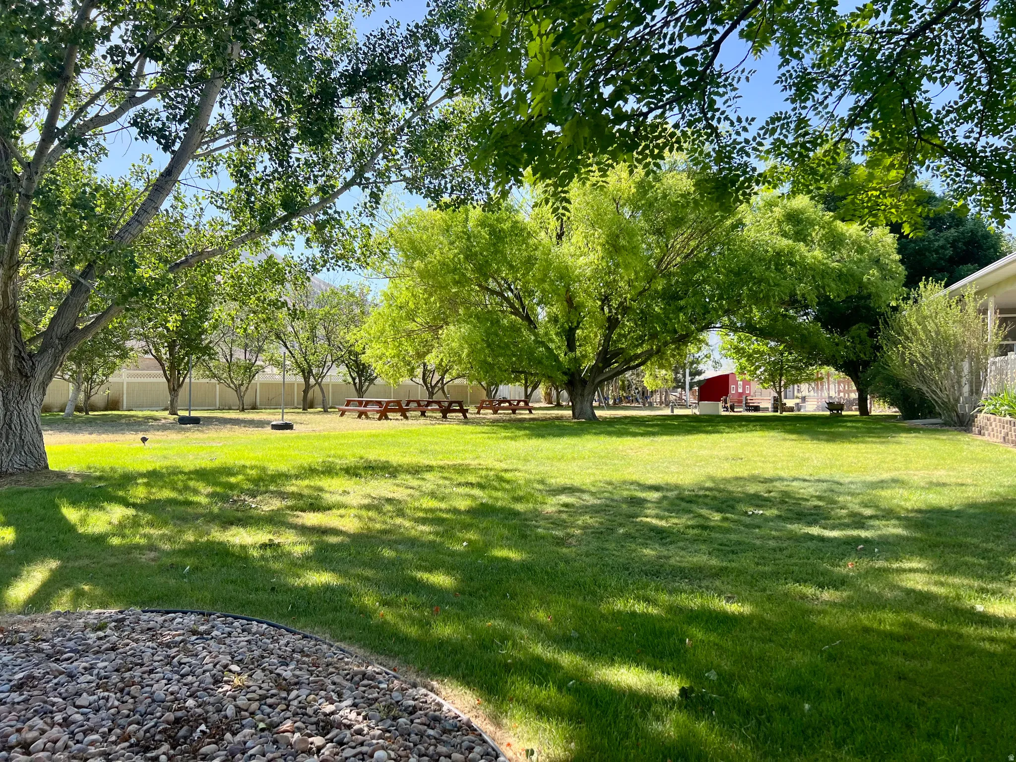 Expansive green lawn framed by mature trees, featuring multiple picnic tables and a white perimeter fence