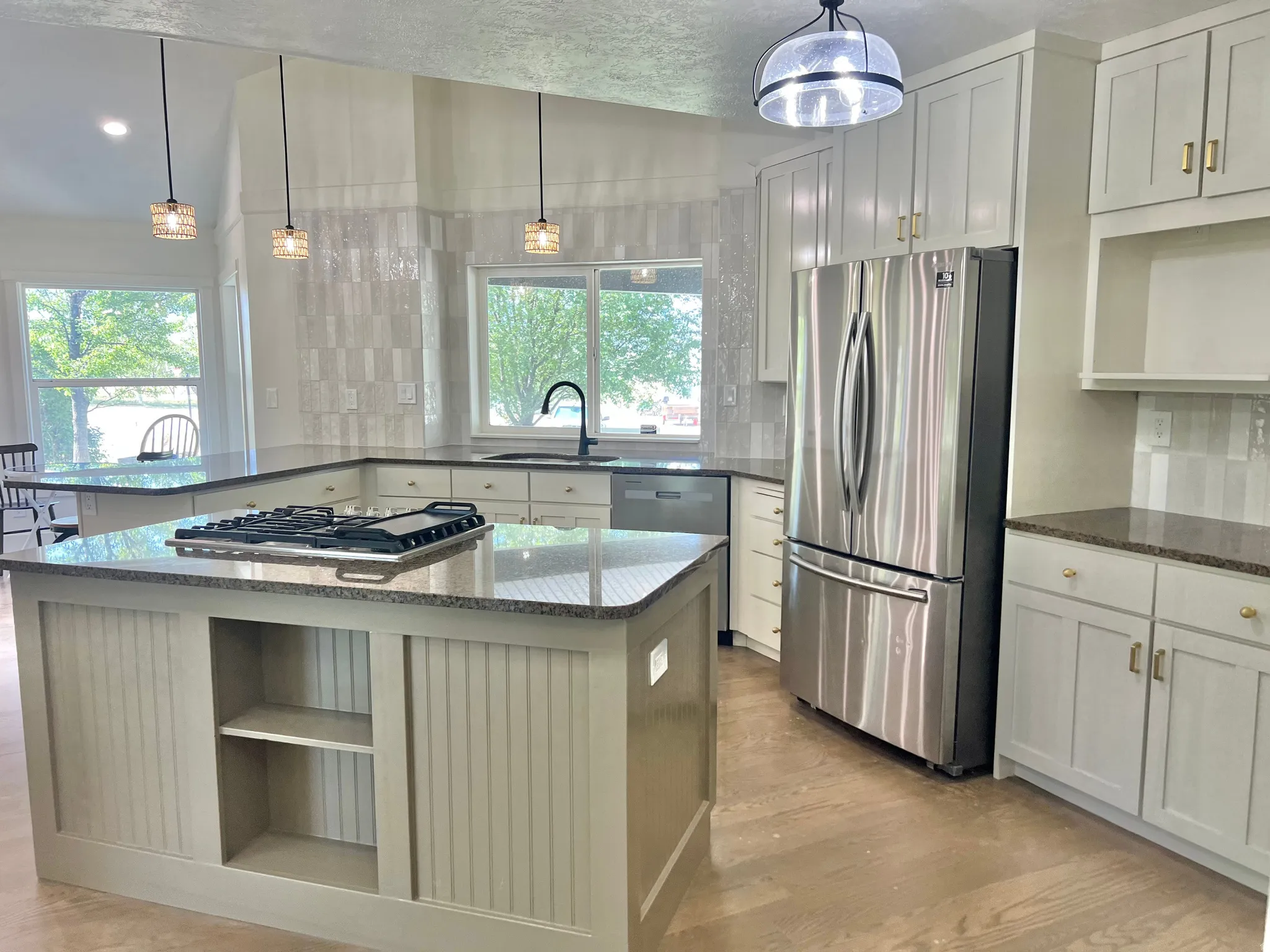 Bright kitchen featuring a central island with an integrated cooktop, granite countertops, stainless steel appliances, white shaker cabinetry with gold hardware, and wood-finish flooring