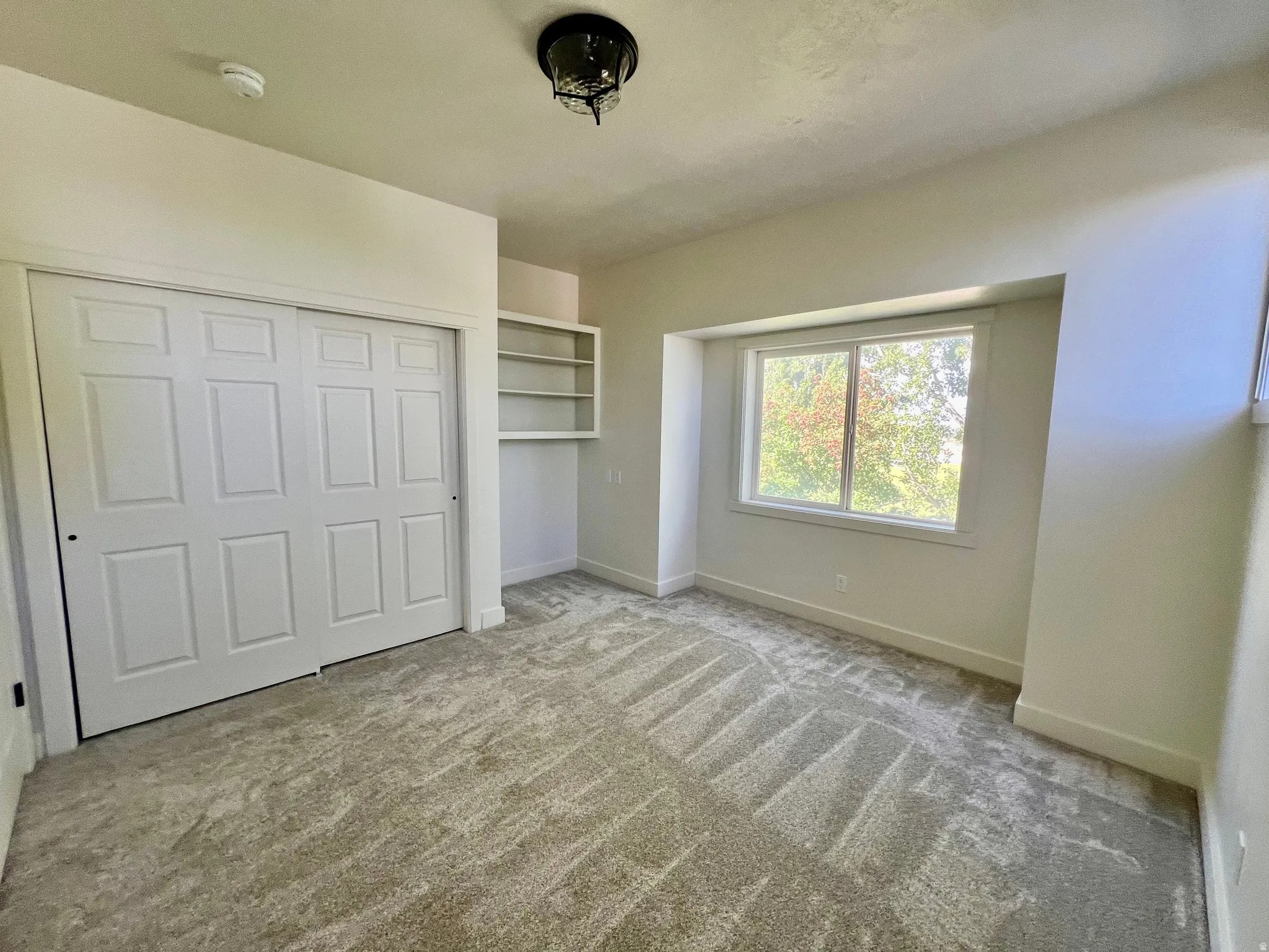 Bright room featuring a window with exterior scenery, light-toned carpet flooring, white bi-fold closet doors, built-in shelving, and a ceiling-mounted light fixture