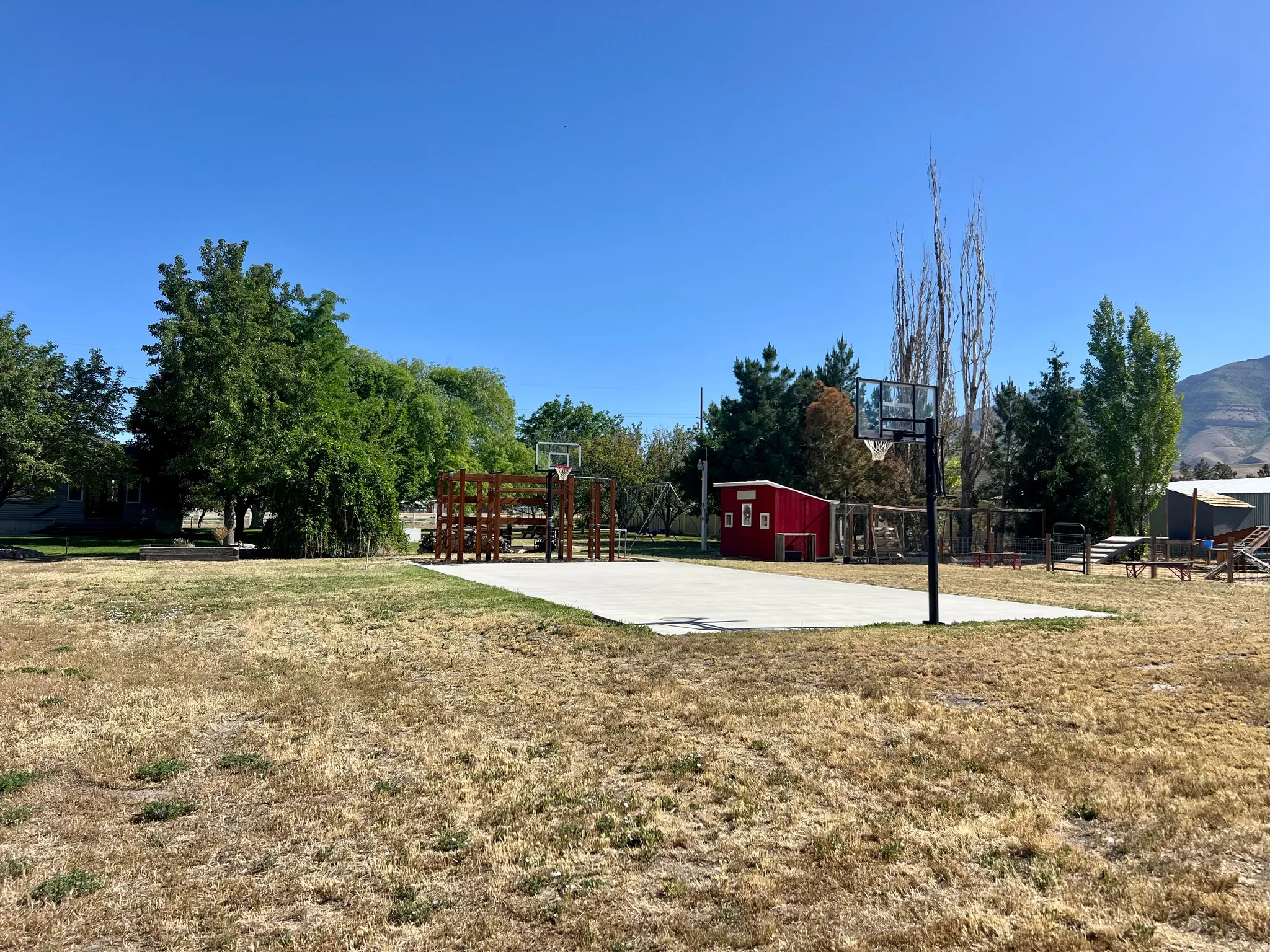 Expansive outdoor space featuring a concrete sports court with two basketball hoops, a red storage shed, mature trees, and a wooden play structure