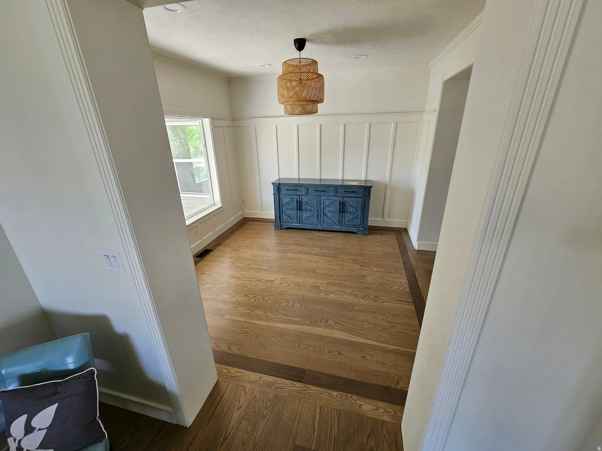 Spacious room featuring wood-finish flooring with a contrasting border, board and batten wall paneling, a large window, and a woven pendant light fixture