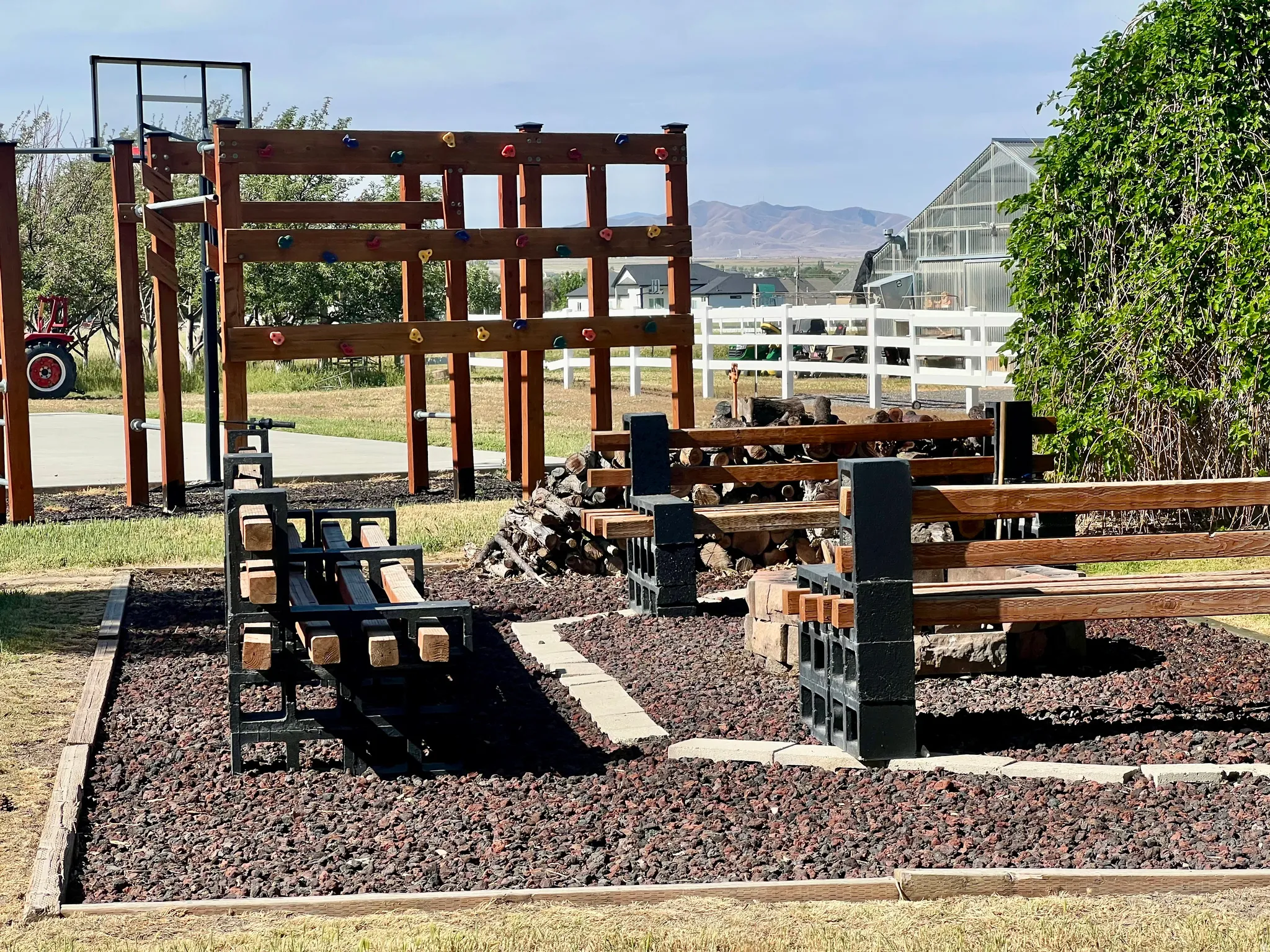 Outdoor recreational area featuring a wooden climbing structure with colorful holds, a basketball hoop, and two dark-toned metal and wood benches