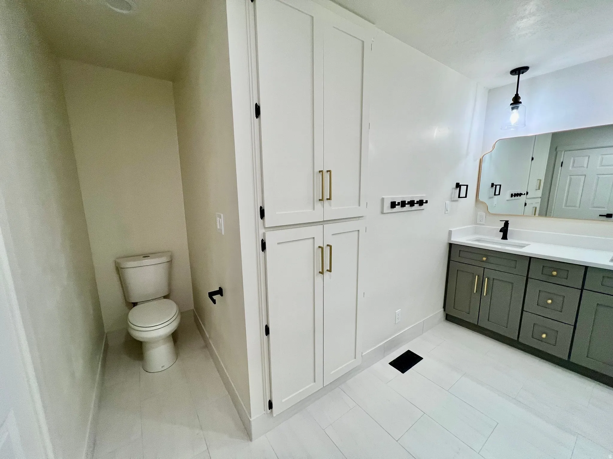 Bathroom featuring white tile flooring, a dual-sink vanity with gray cabinetry and gold-tone hardware, a decorative mirror, and an adjacent built-in storage cabinet with gold-tone hardware