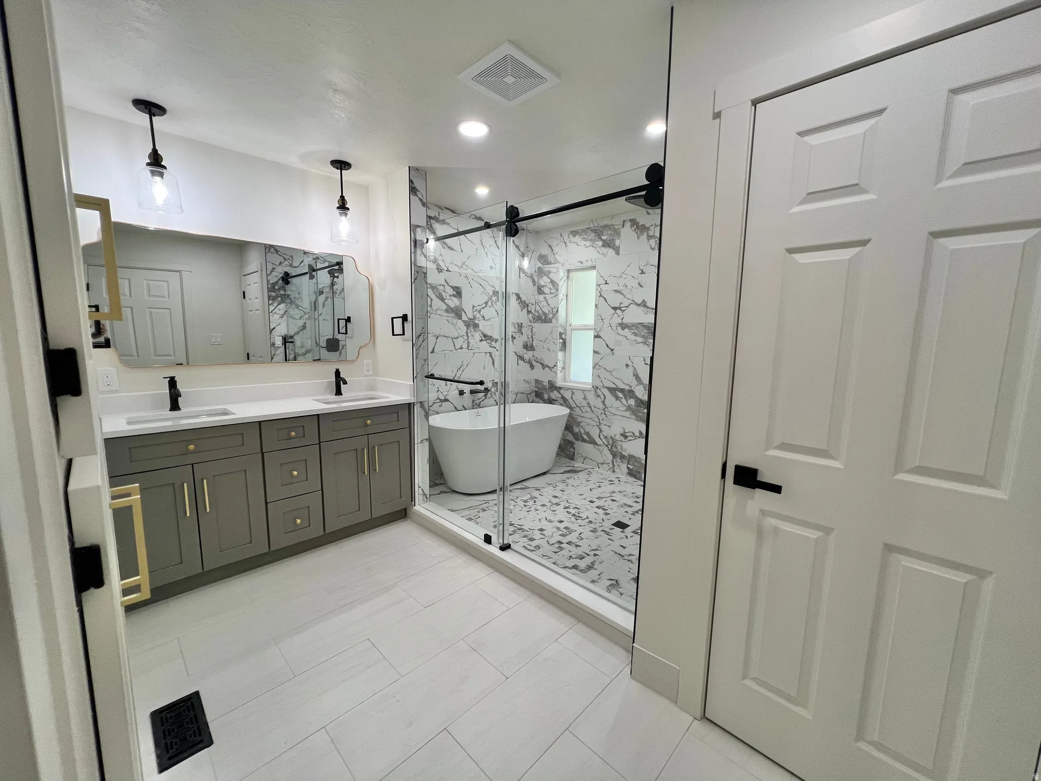 Renovated bathroom featuring a dual vanity with grey cabinetry and gold-tone hardware, a freestanding soaking tub, and an enclosed shower with full-height marble-patterned tile