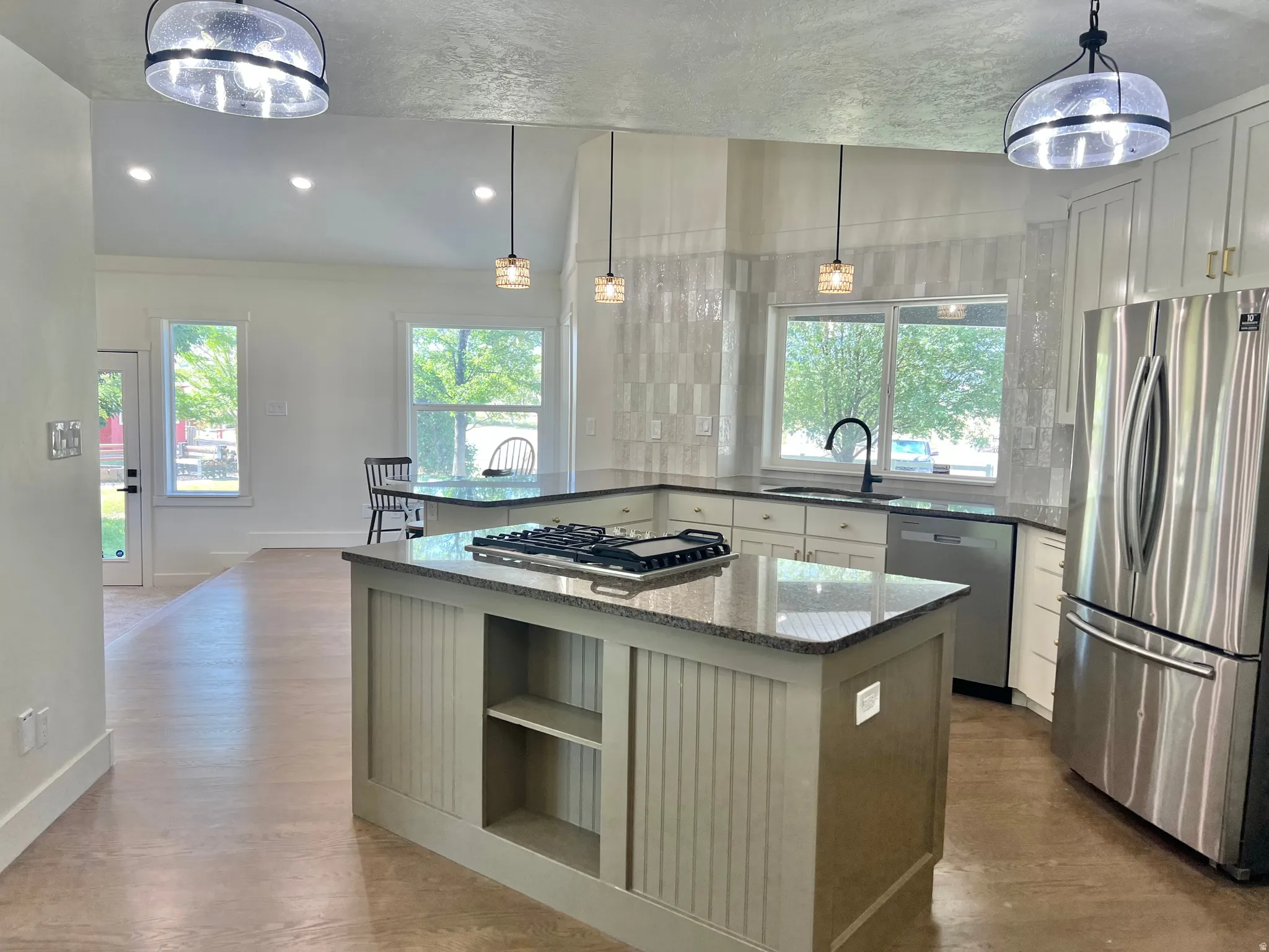 Open-concept kitchen featuring an island with a built-in gas cooktop, granite countertops, stainless steel appliances, white cabinetry with gold-tone hardware, and a tile backsplash