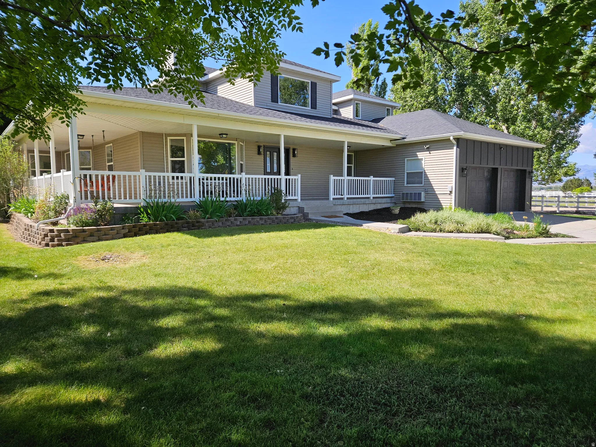 Two-story residence featuring a full-width covered front porch with white railings, multiple dormer windows, an attached two-car garage with black doors, and a stone-block retaining wall with landscaping
