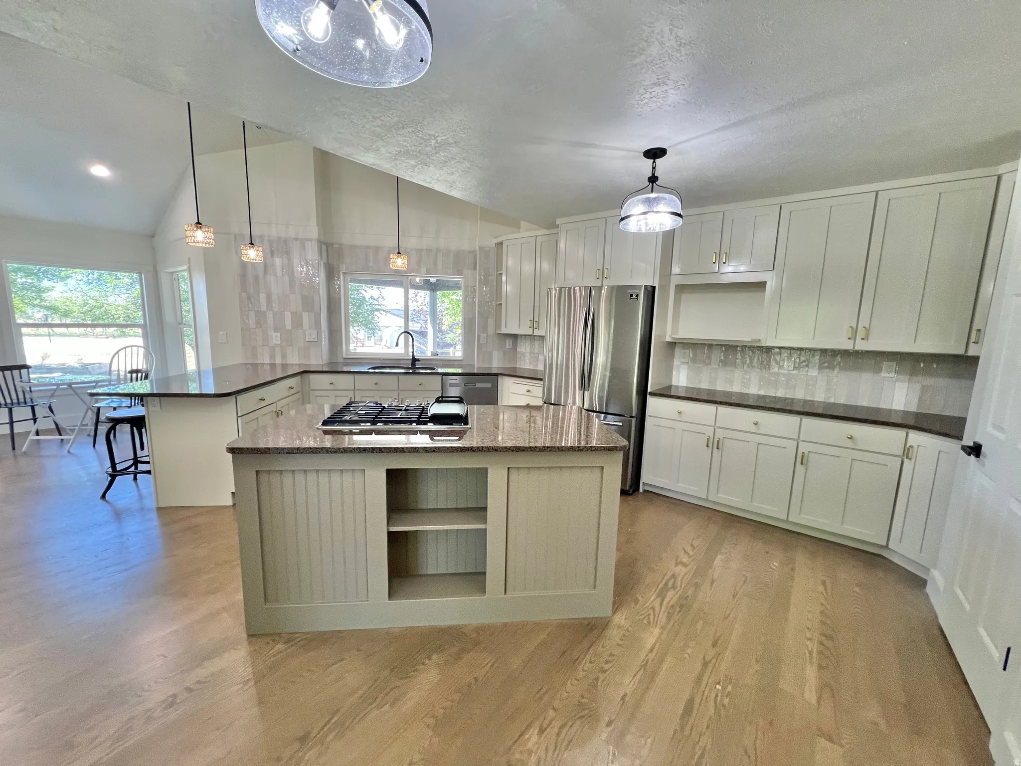 Open-concept kitchen featuring an expansive center island with a built-in cooktop, light-toned wood-finish flooring, white cabinetry with gold-tone hardware, and a tile backsplash