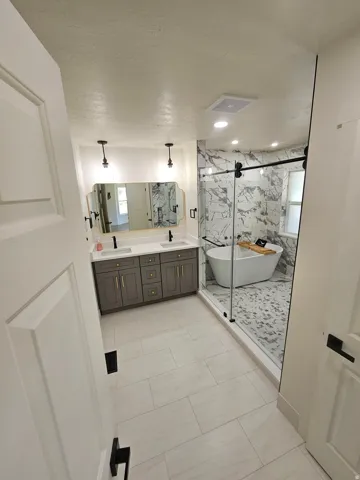 Modern bathroom featuring a dual vanity with gray cabinetry, a freestanding soaking tub, a walk-in shower with a glass enclosure, marble-look tile, and large format floor tiling
