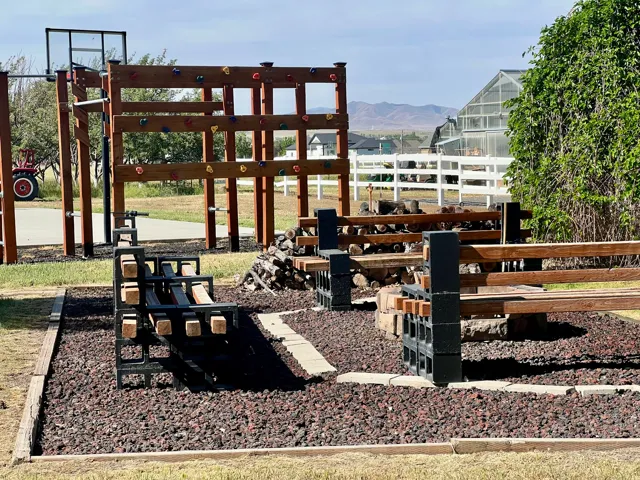 Outdoor recreational area featuring a wooden climbing structure with colorful holds, a basketball hoop, and two dark-toned metal and wood benches