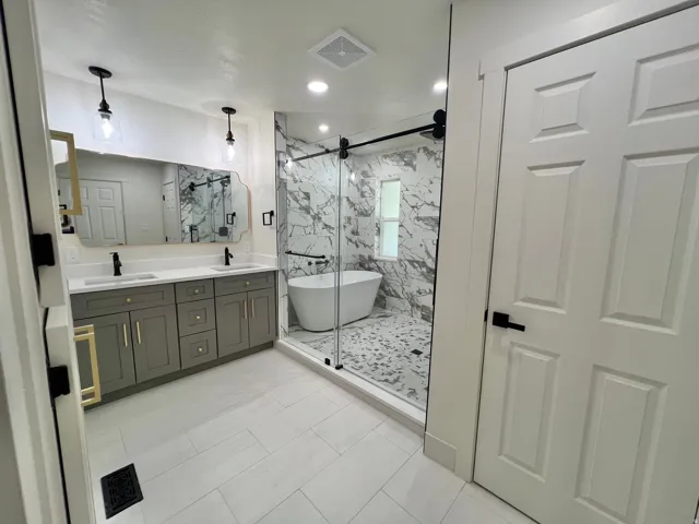 Renovated bathroom featuring a dual vanity with grey cabinetry and gold-tone hardware, a freestanding soaking tub, and an enclosed shower with full-height marble-patterned tile