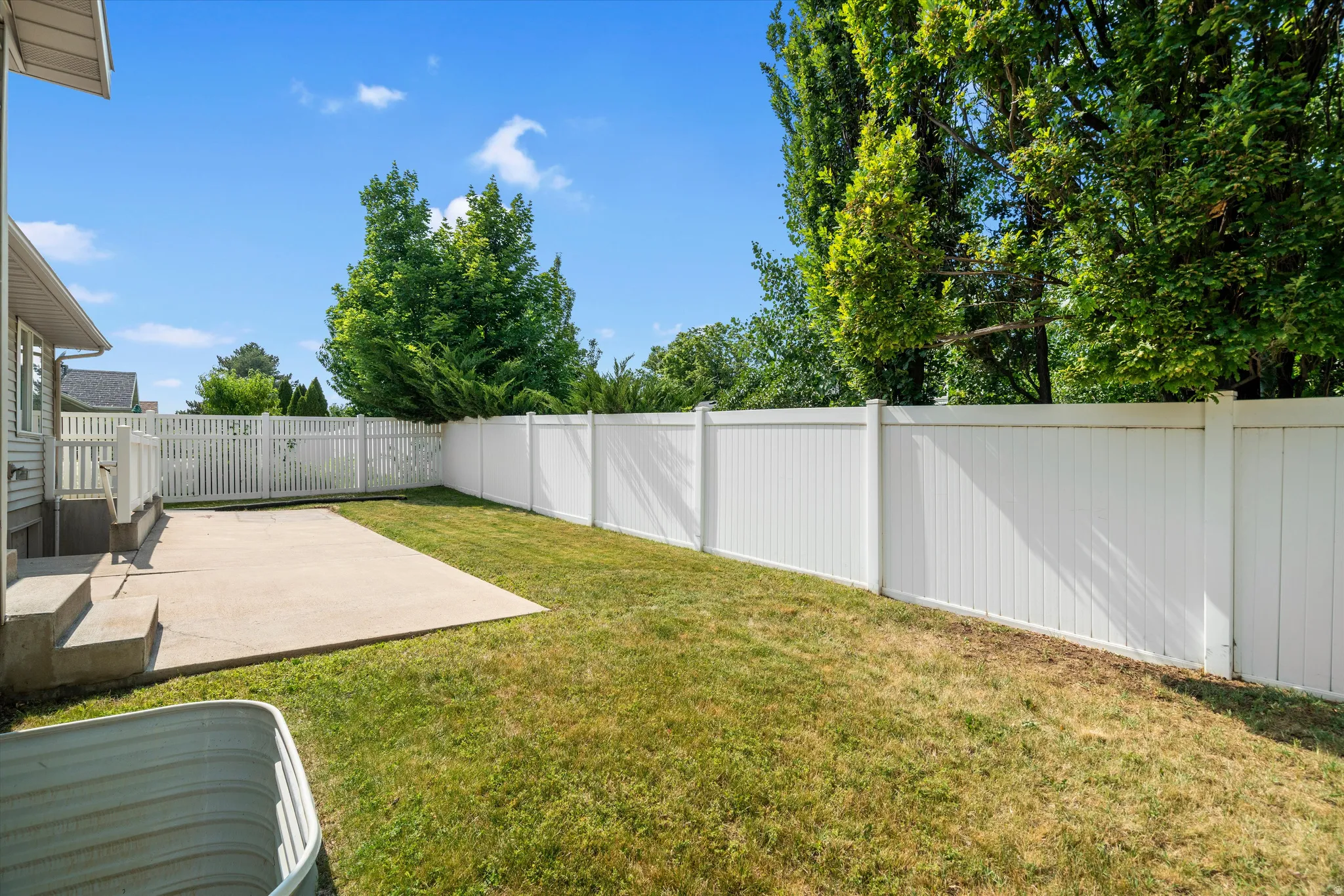 Expansive backyard featuring a concrete patio, a full white vinyl privacy fence, and mature trees surrounding the perimeter